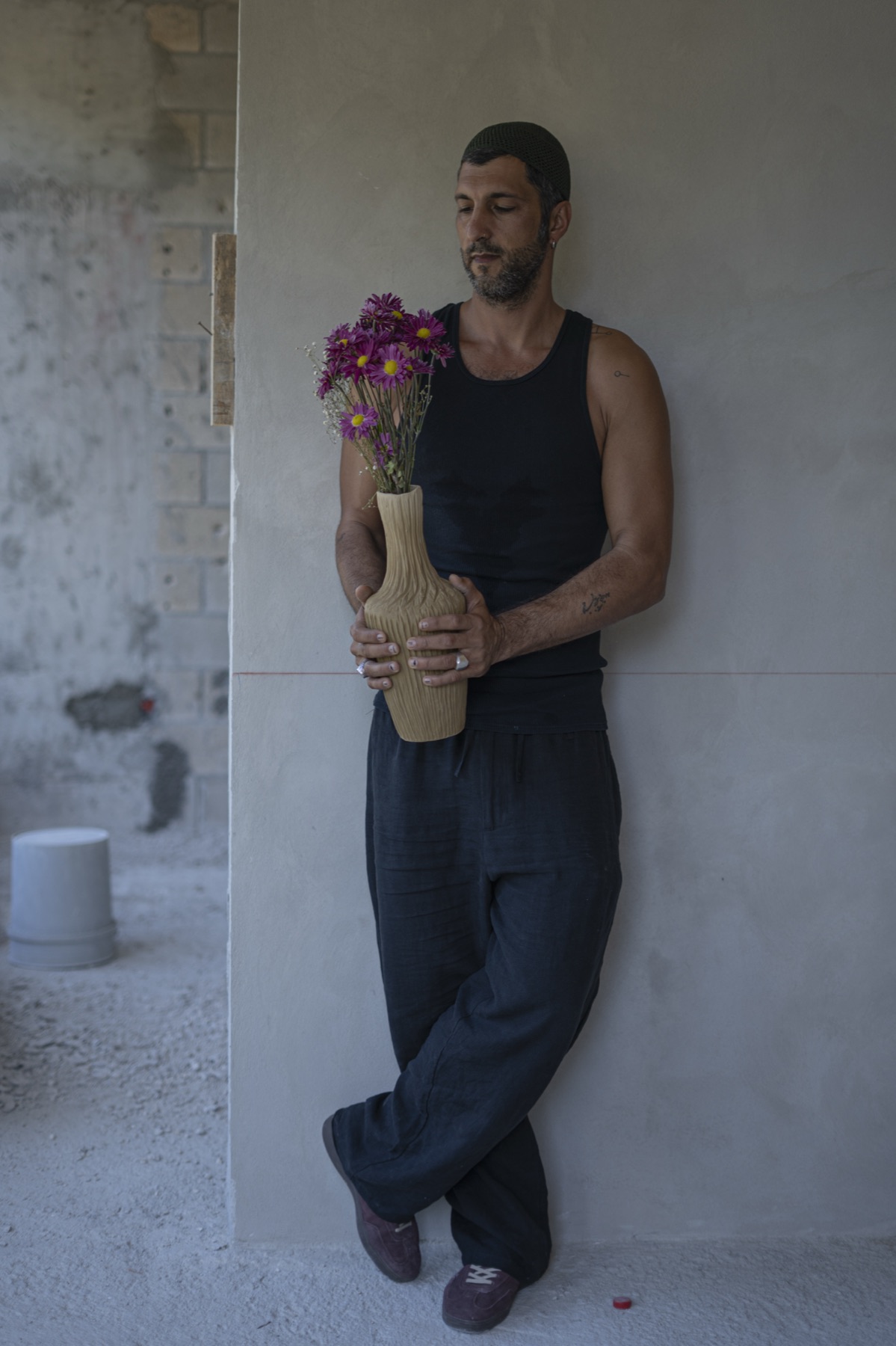 The maker against a plaster wall, holding a vessel filled with pink flowers.