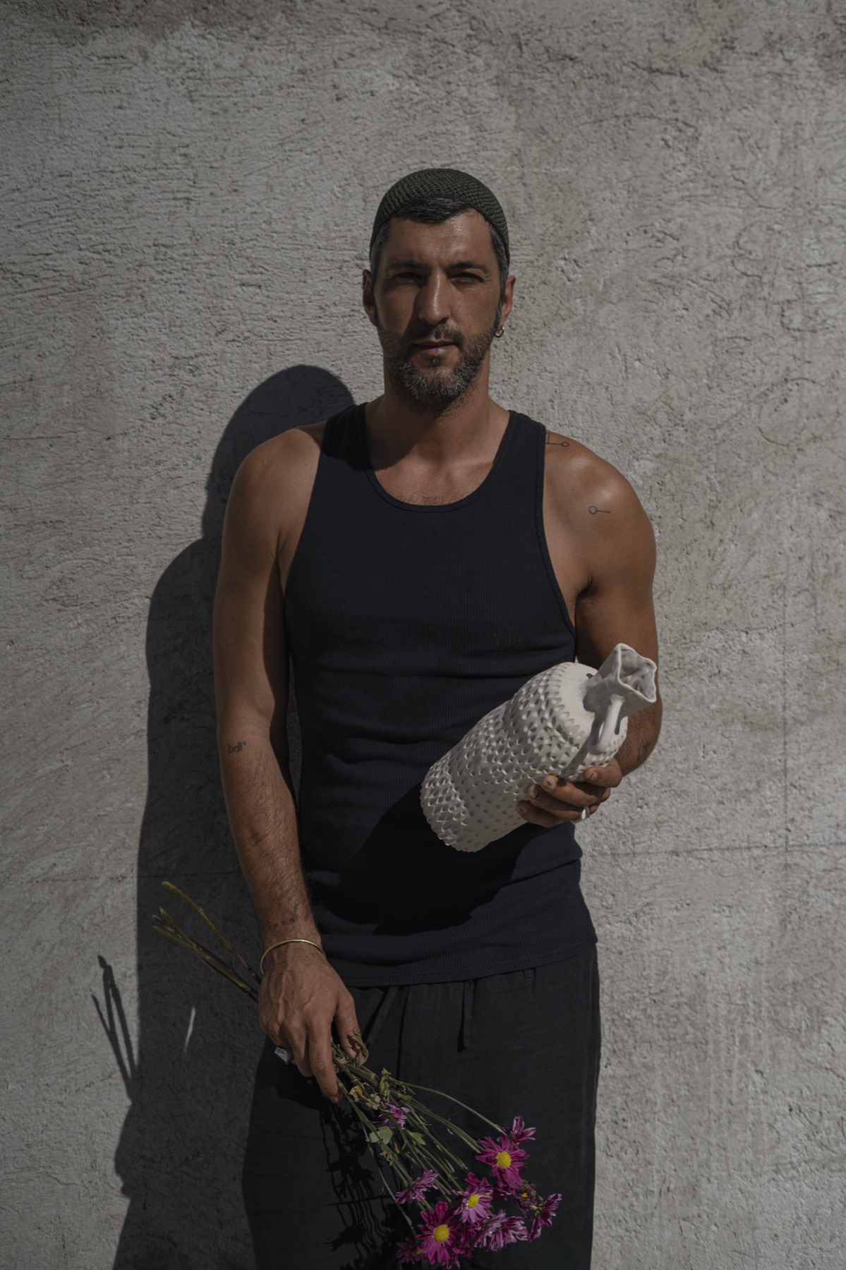 The maker standing against plaster, a white vessel in one hand and cut flowers in the other.