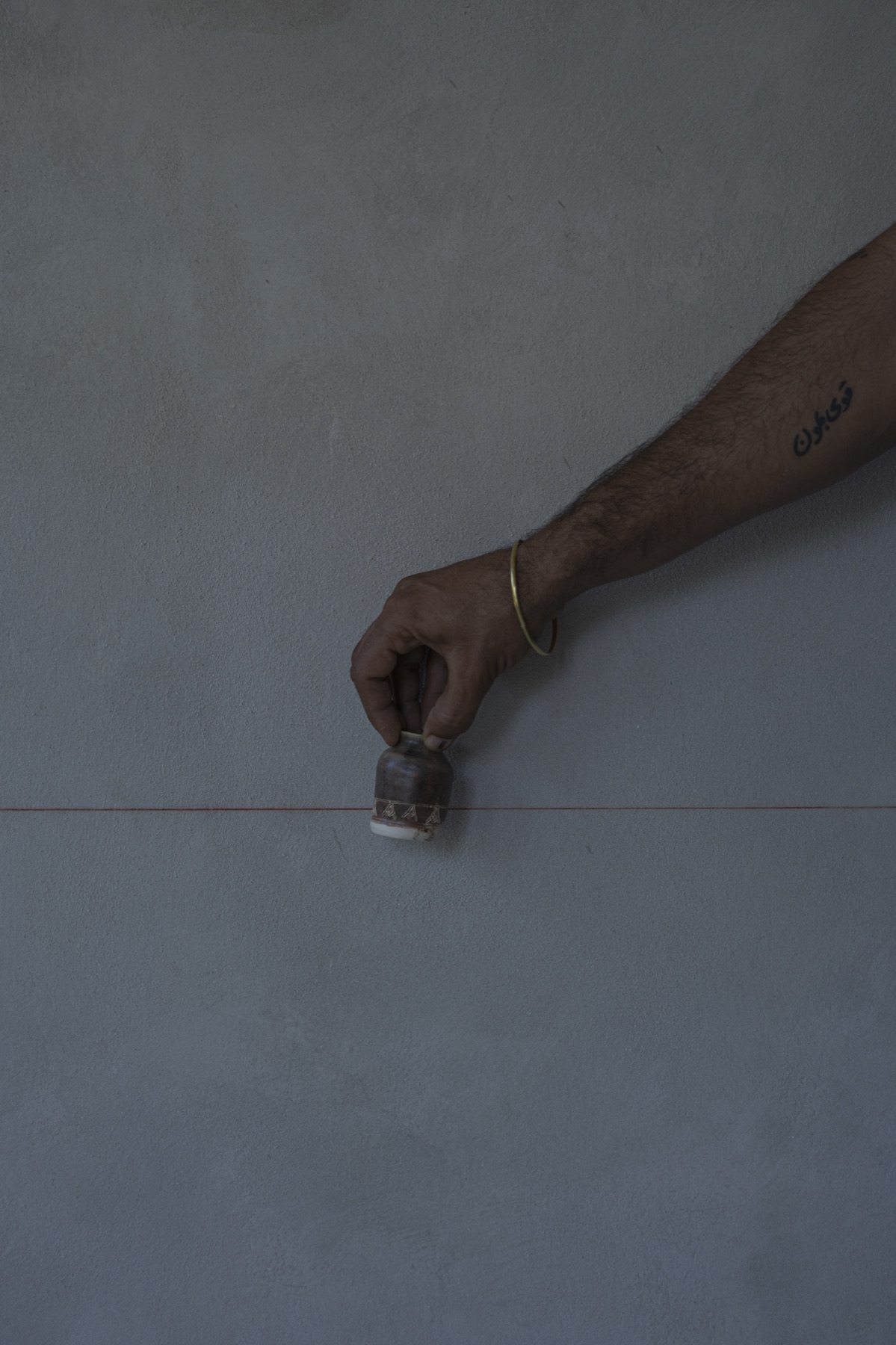 A tattooed forearm setting a small vessel down against a bare plaster wall.