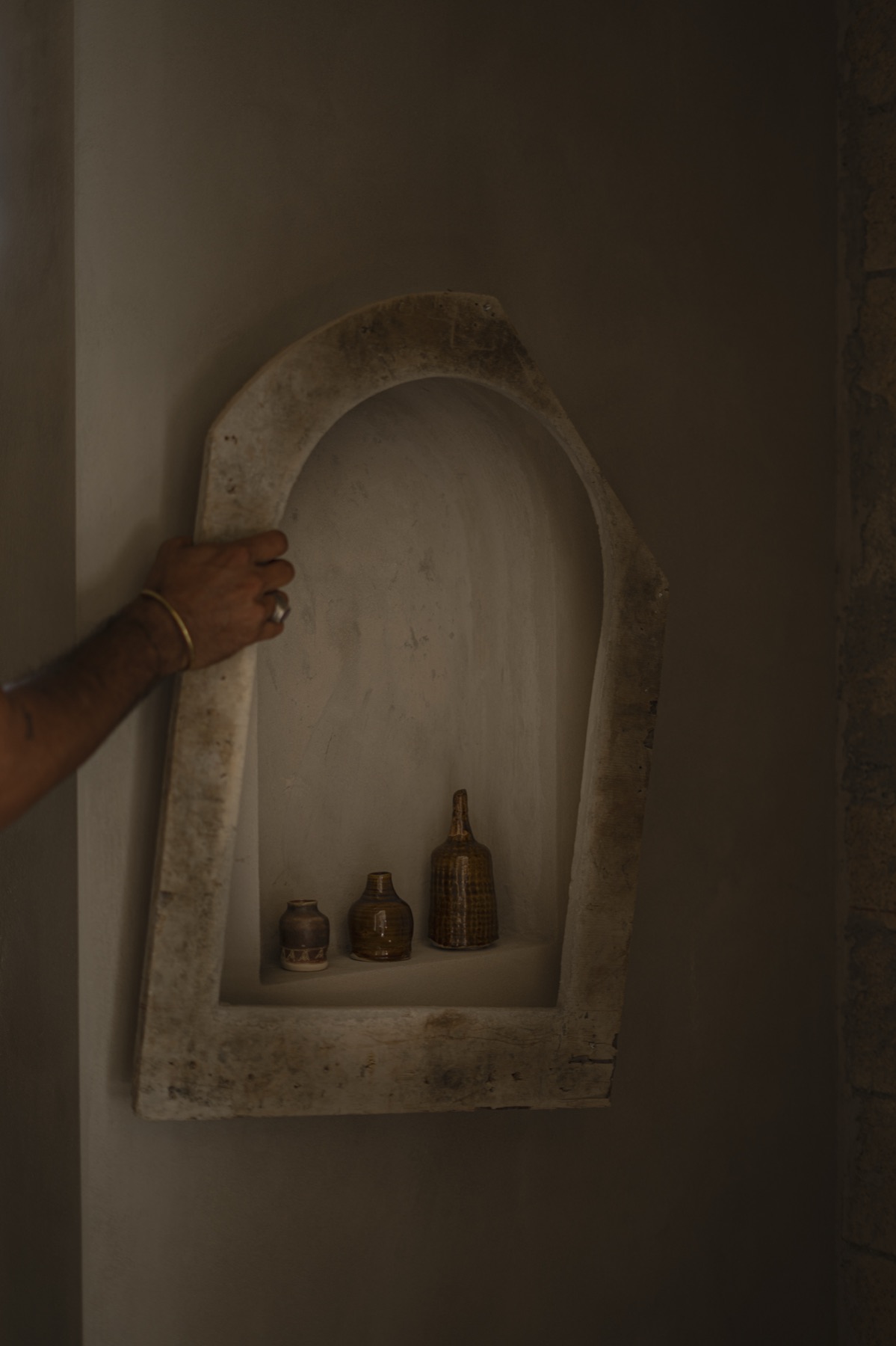 A hand reaching into an arched plaster niche holding small dark bottles.