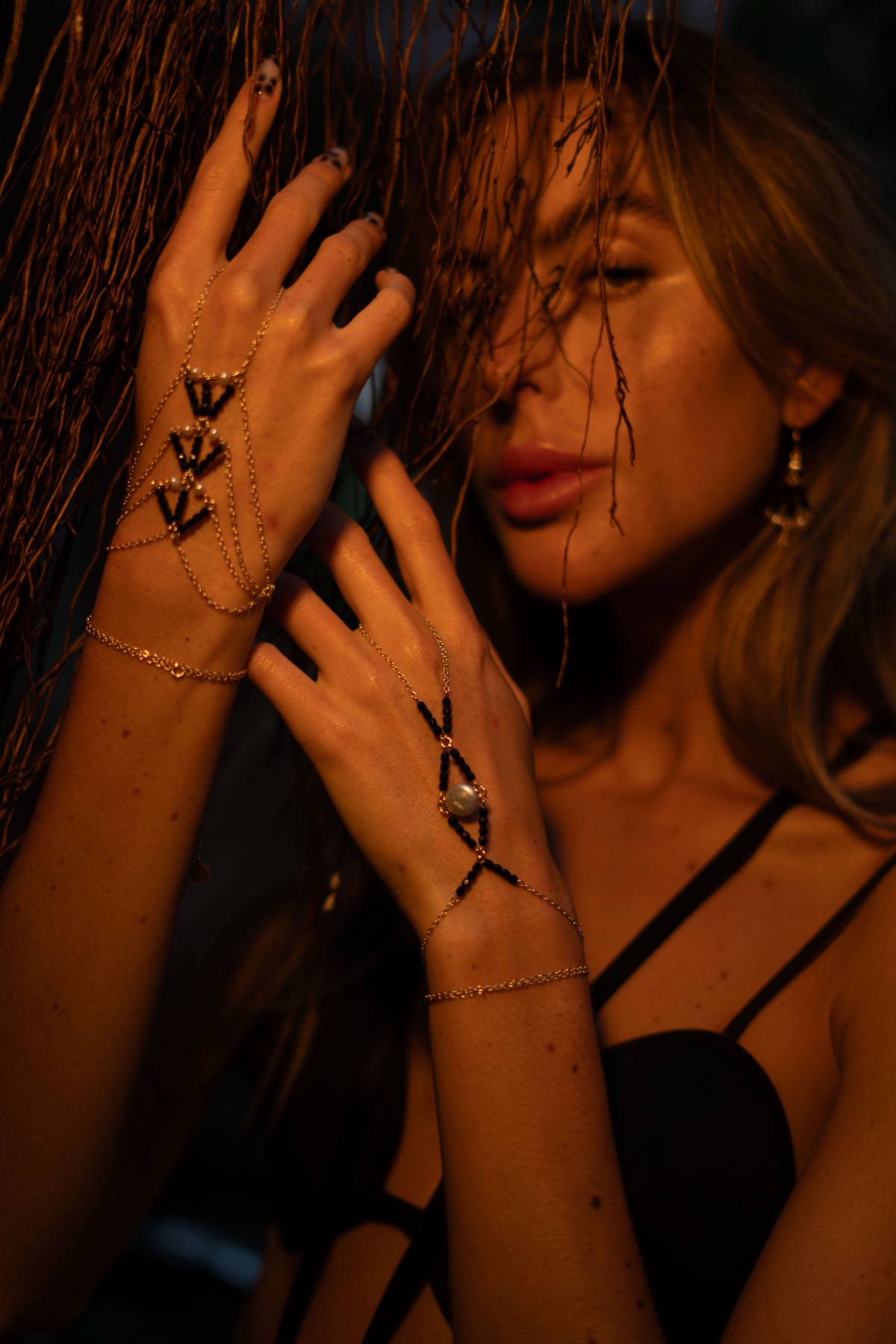 Two hands against hanging roots wear gold hand chains set with black stones and a baroque pearl.
