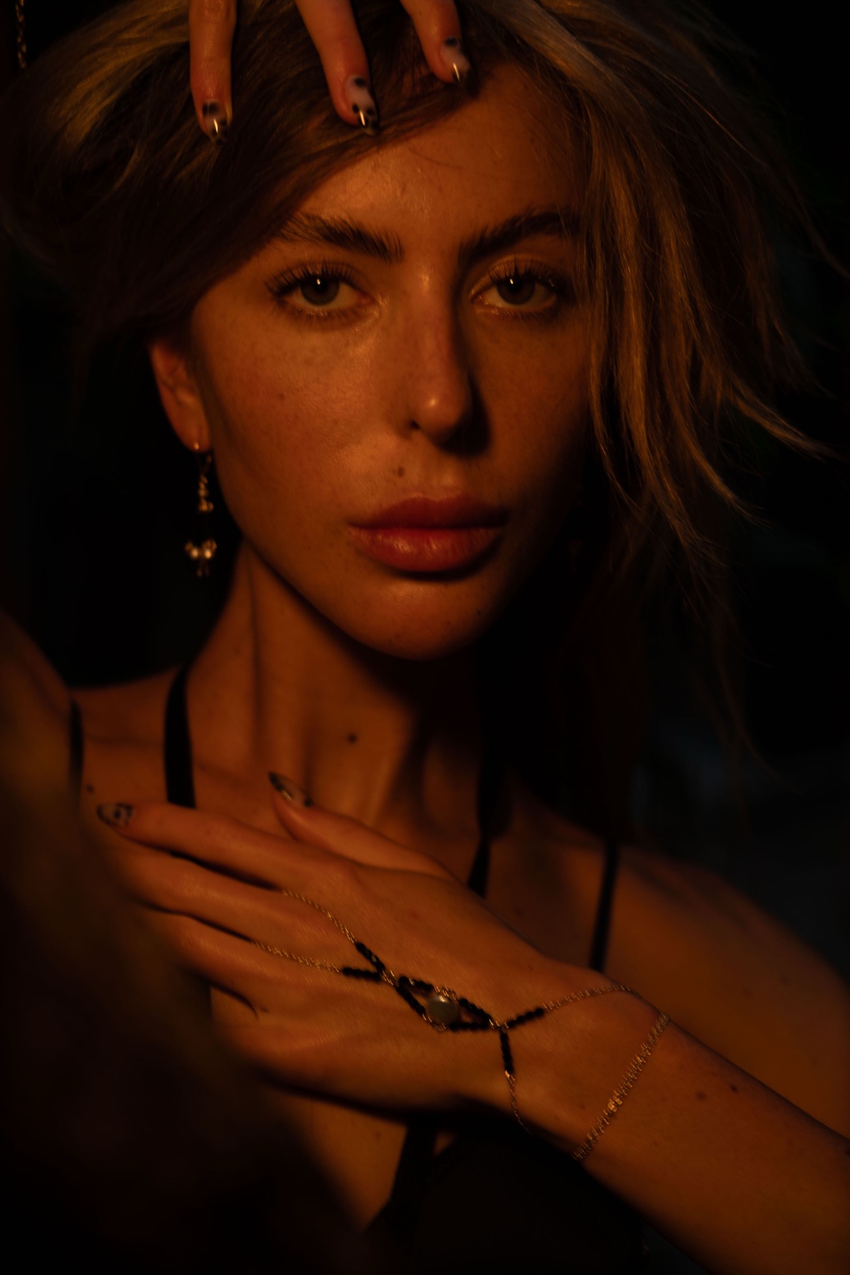 A woman looks straight to camera, one hand in her hair, a gold and black hand chain across her fingers.