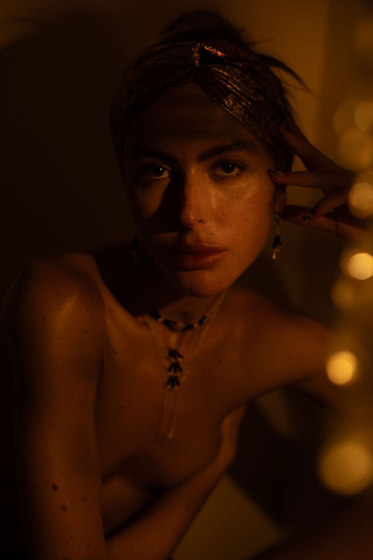 A woman in a gold brocade headwrap looks to camera wearing a fine gold and black chain necklace.