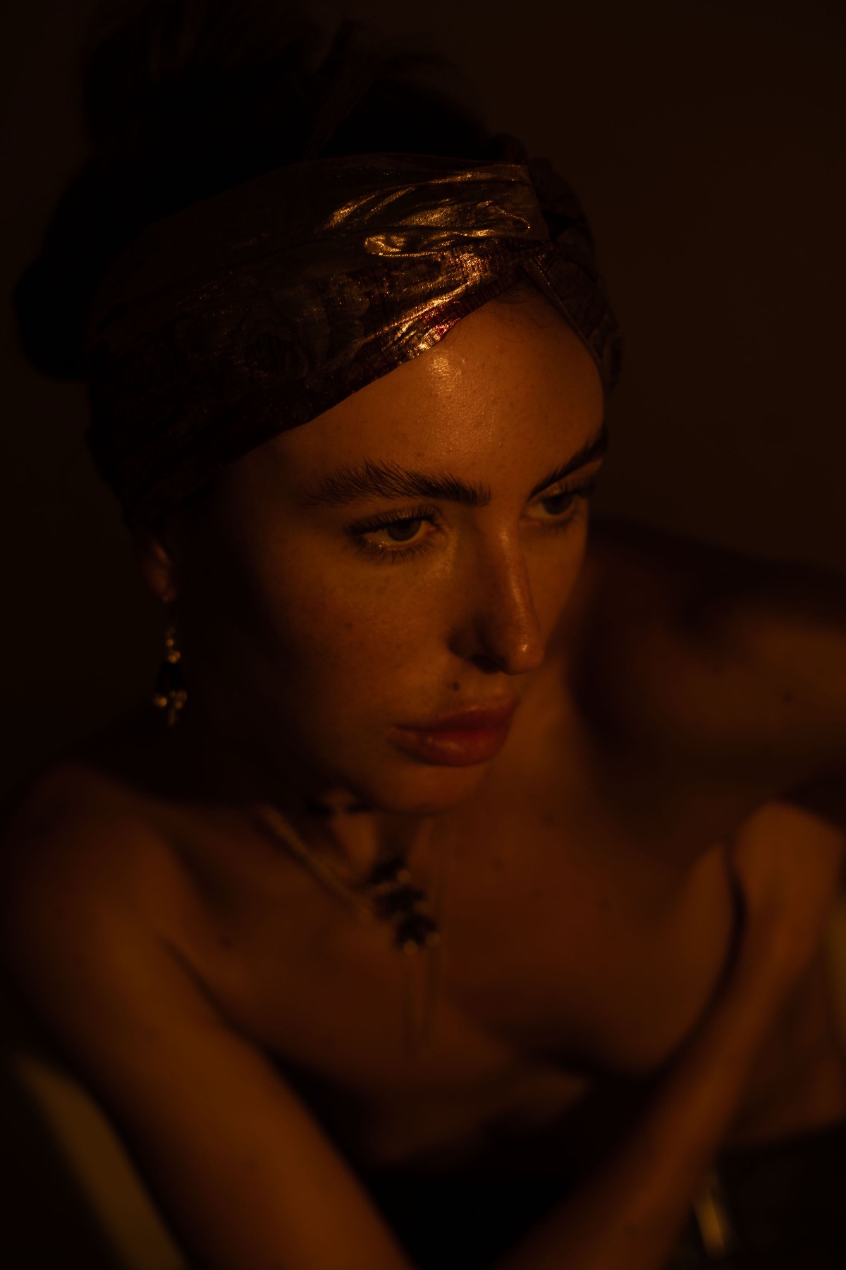 Close portrait of a woman in a bronze brocade headwrap with a gold drop earring, warm shadow behind.