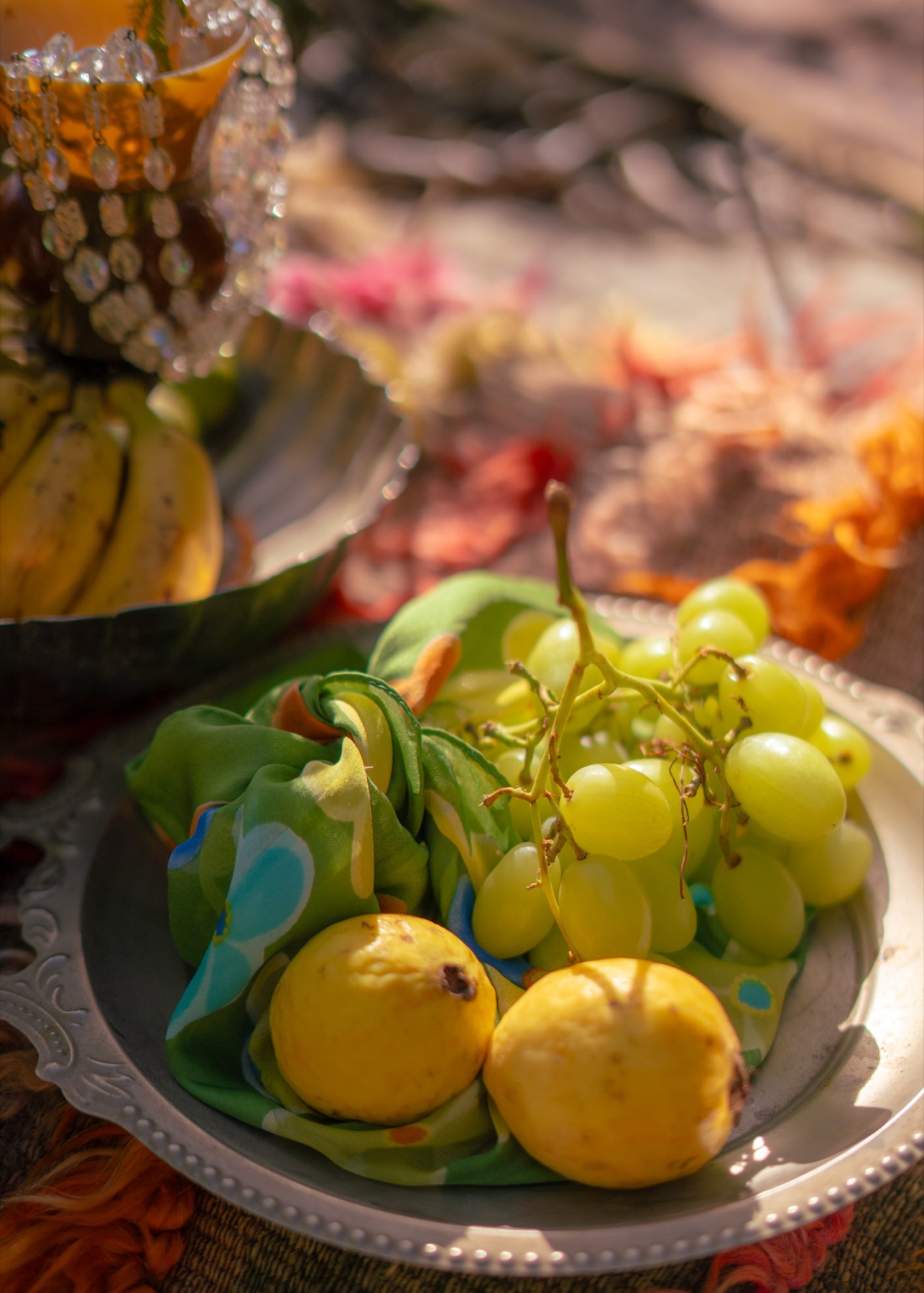 Grapes, guavas and a printed silk scarf on an engraved silver dish beside a beaded goblet, styled on layered textiles.