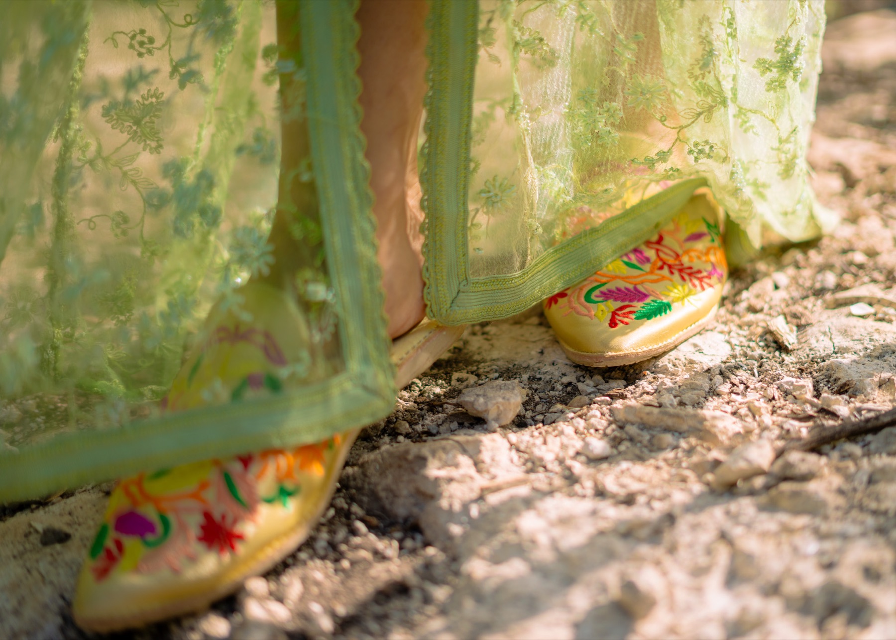 Embroidered flat slippers and bare feet under the sheer green hem of a lace kaftan on a limestone path.