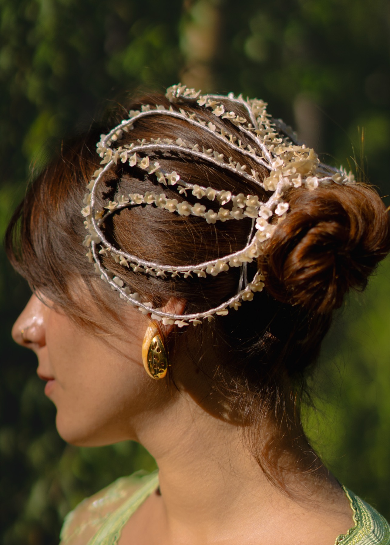 Profile of a woman wearing a beaded floral headpiece, gold teardrop earring and green lace, against jungle foliage.
