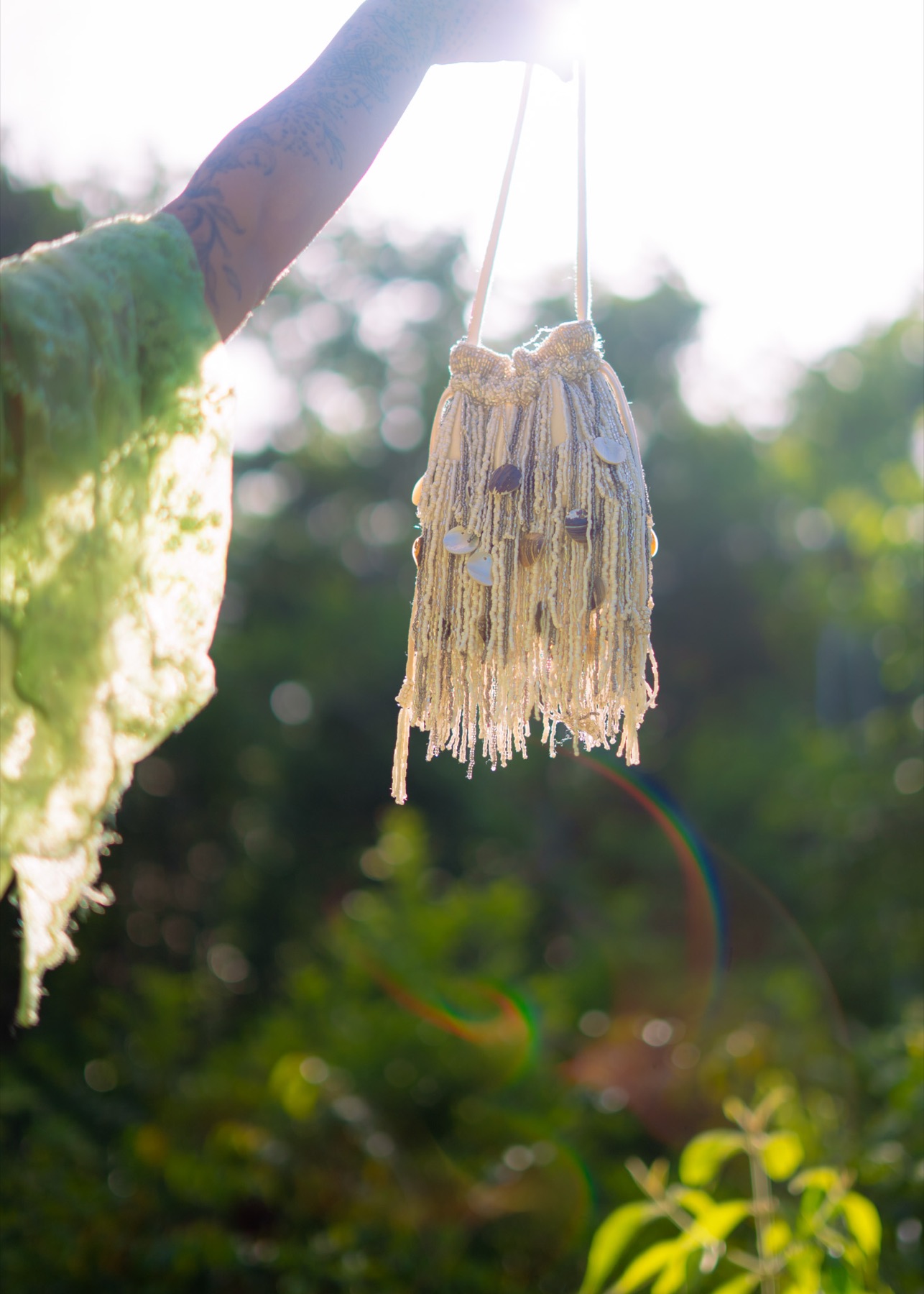 A hand lifts a shell-and-fringe tasselled bag into the sunlight, lens flare across the frame.
