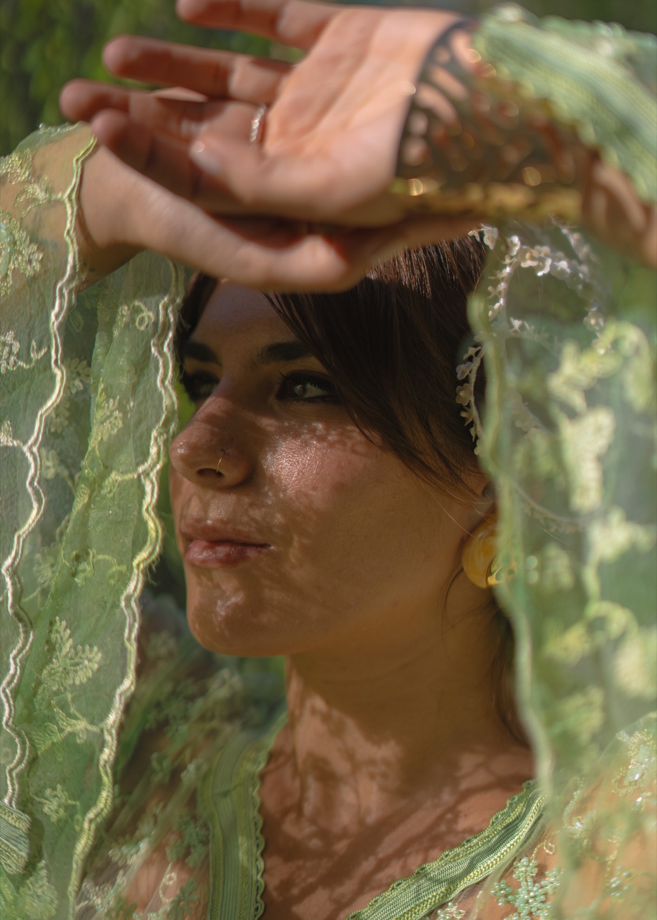 Dappled light falls across a woman's face as she holds a green lace veil above her head.