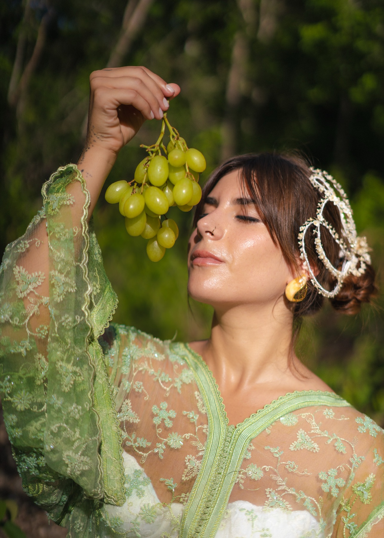 A woman in green lace holds a bunch of green grapes to her mouth, eyes closed, jungle light behind her.