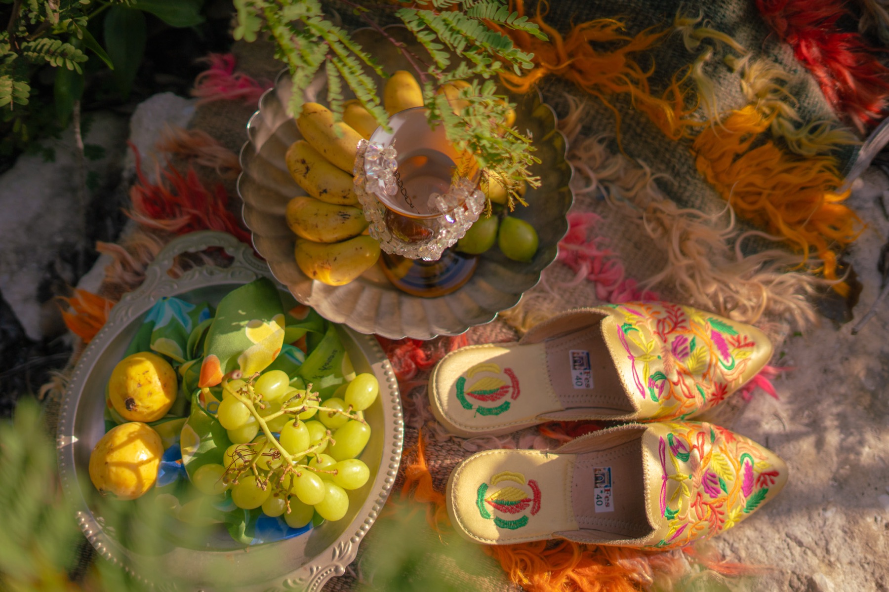 Overhead still life of embroidered slippers, silver dishes of fruit and a beaded goblet on a fringed blanket over limestone.