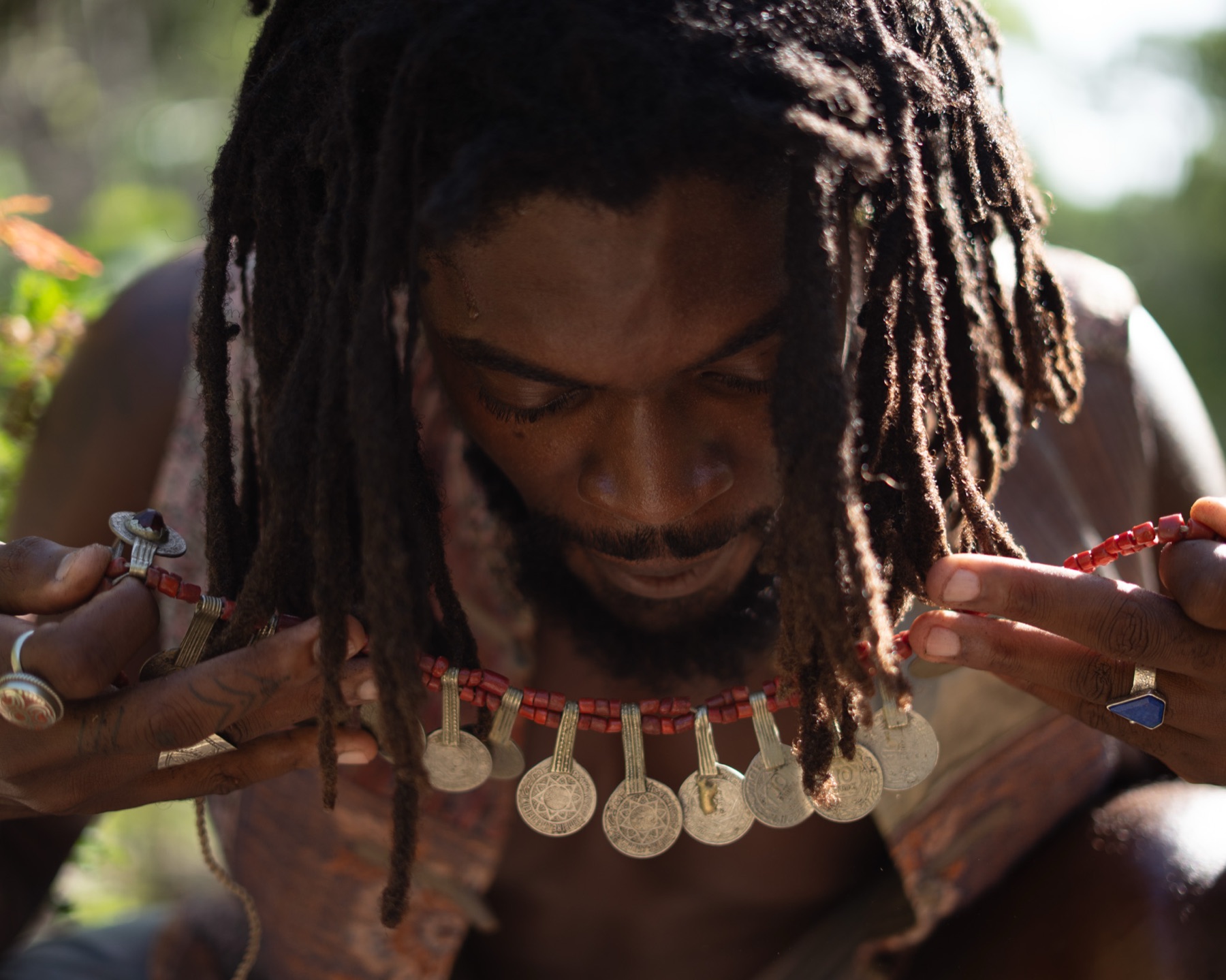 A man with locs lowers his head as he lifts a silver coin-and-coral necklace towards his chest.