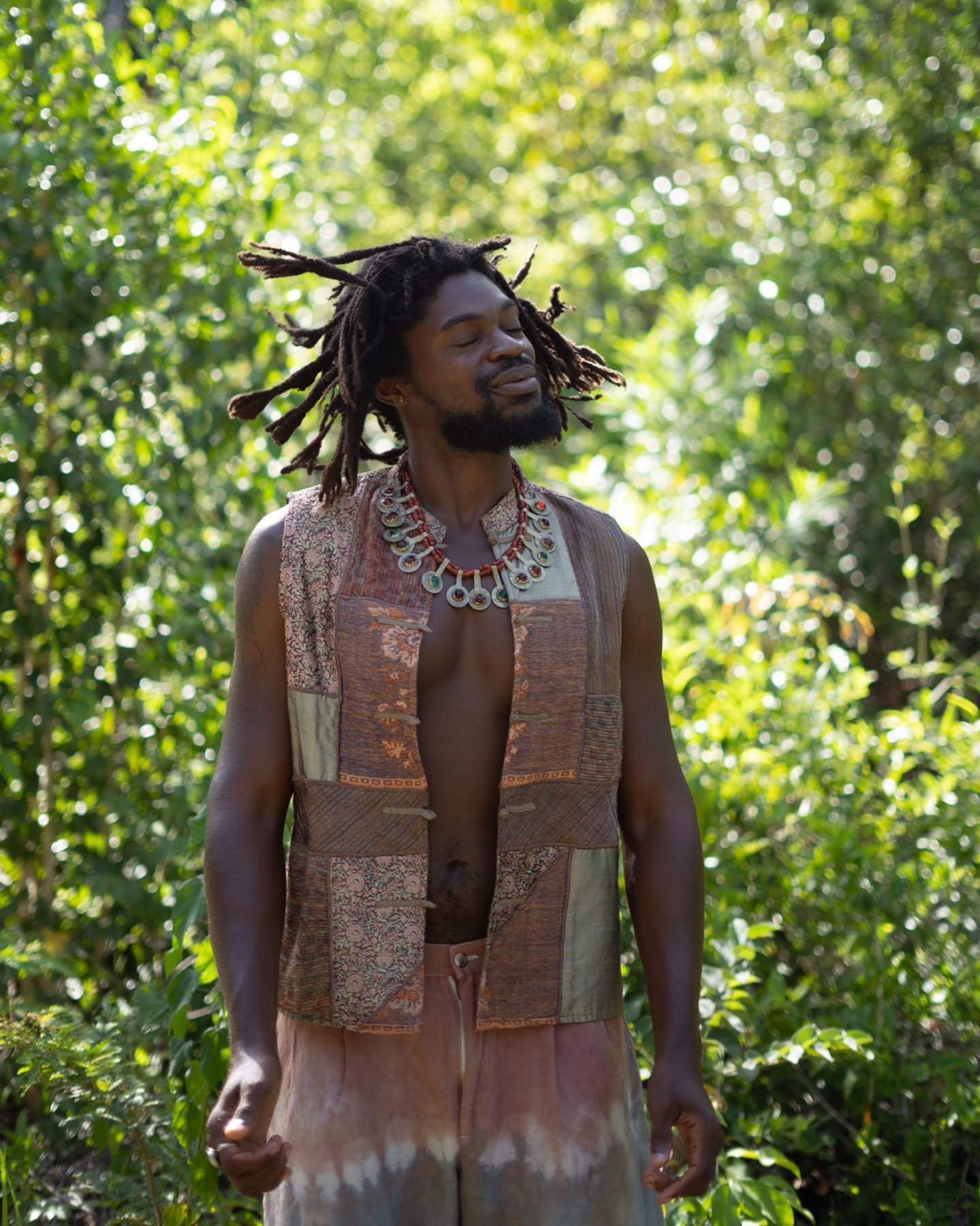 A man in a patchwork waistcoat stands in the jungle wearing a silver and coral talisman necklace, eyes closed in the sun.