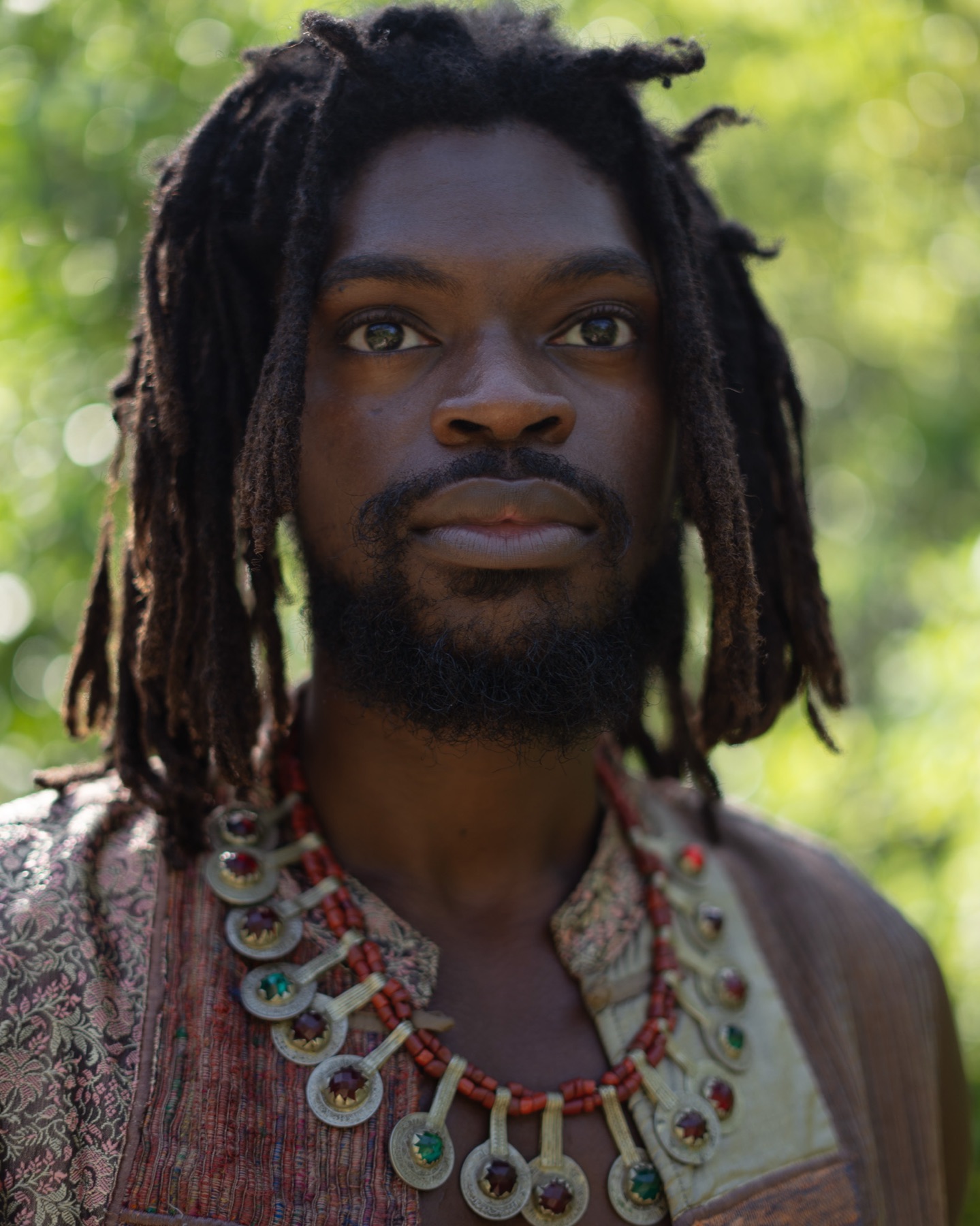 Frontal portrait of a man with locs wearing layered silver and coral necklaces over an embroidered waistcoat.