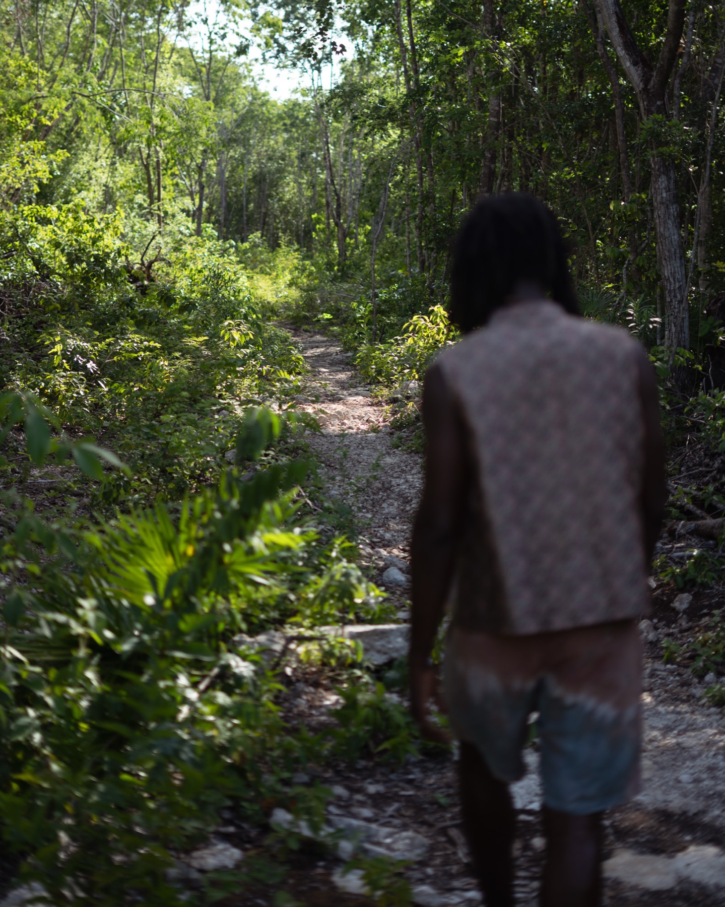 A man in a printed waistcoat walks away down a narrow limestone path into thick Riviera Maya jungle.