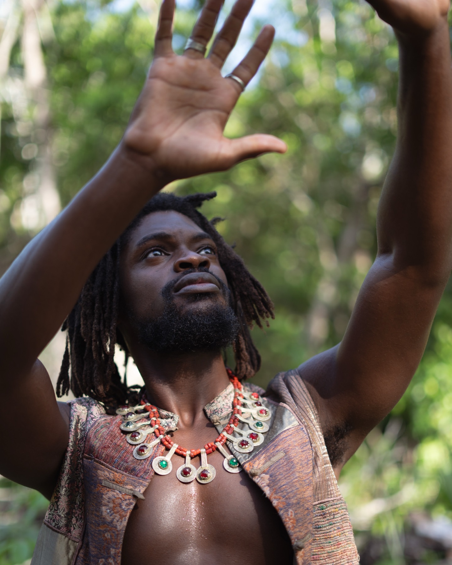 A man raises both arms towards the sky wearing a coral and silver necklace, jungle canopy behind him.