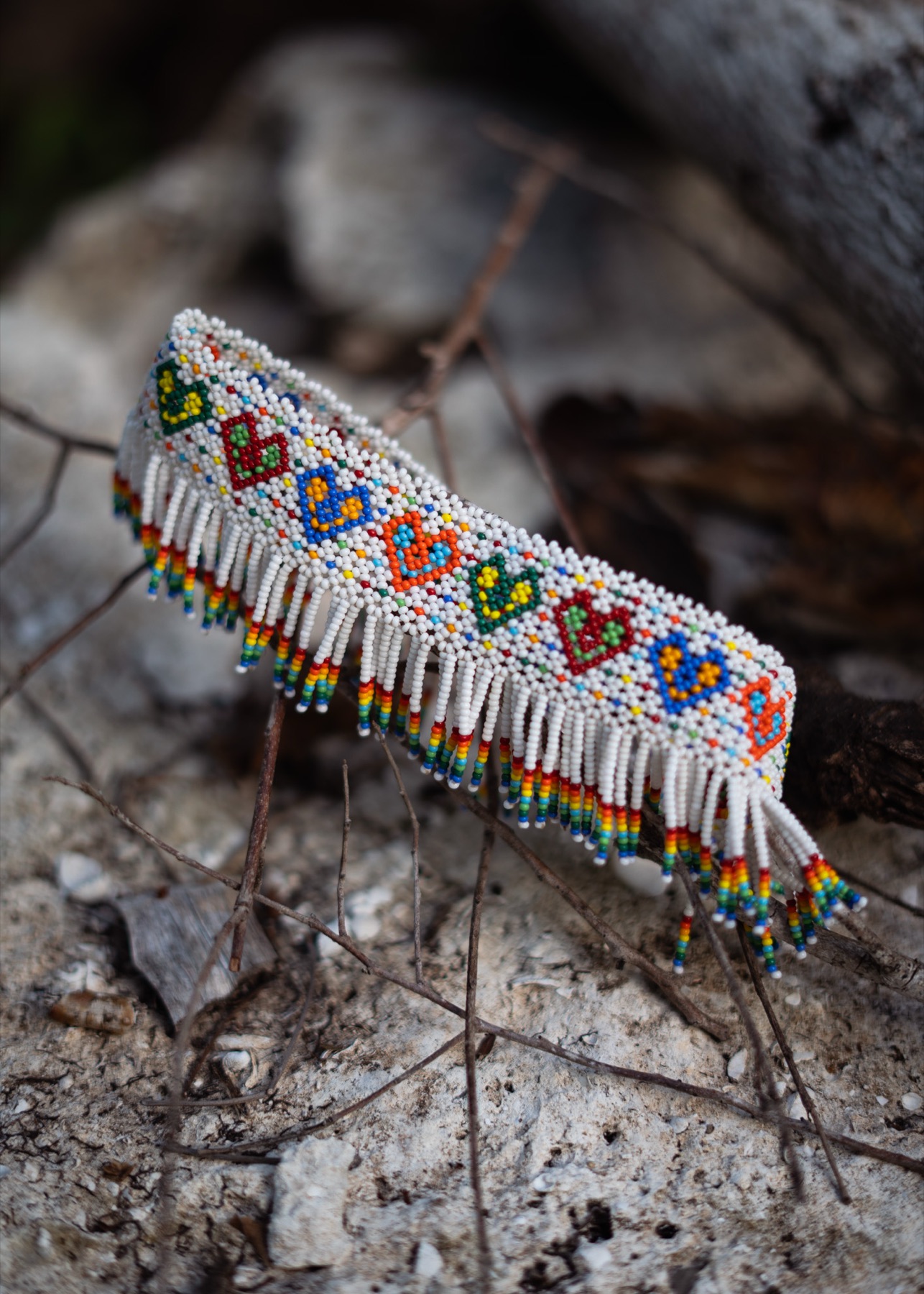 A beaded cuff worked in rainbow geometric patterns with a white fringe, propped on limestone rock.