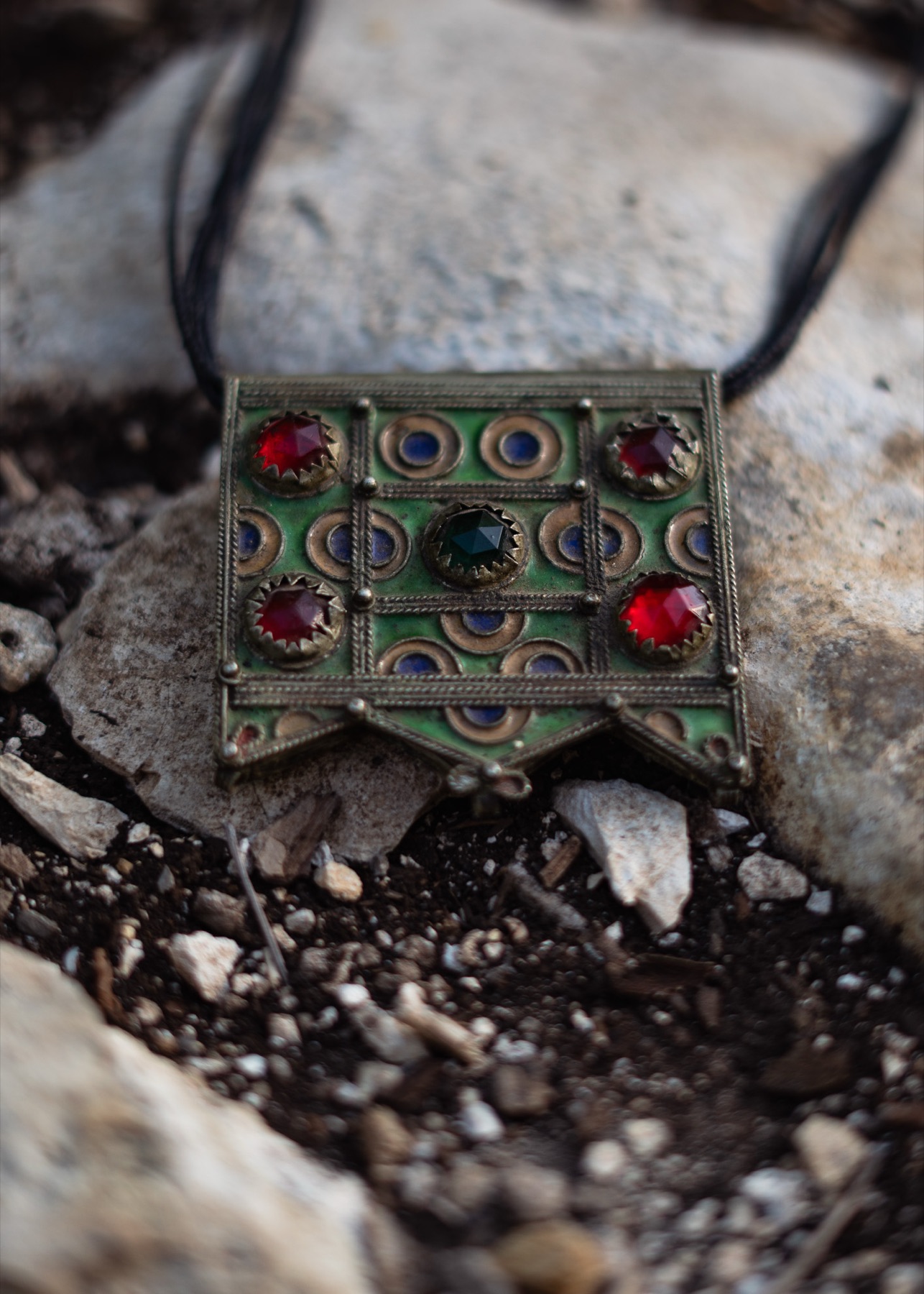 A green enamelled pendant set with red and blue stones, resting on pale stone and dark earth.