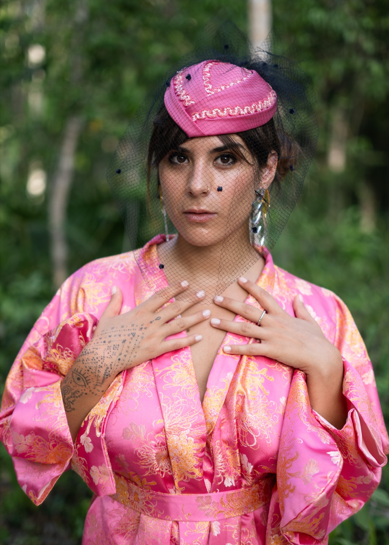 A woman in a veiled pink cap rests both tattooed hands on her chest above a pink and gold brocade kimono.