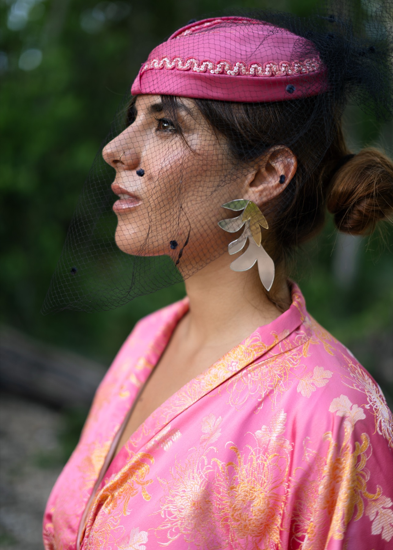 Profile of a woman in a pink veiled cap wearing a long silver leaf earring, jungle bokeh behind.