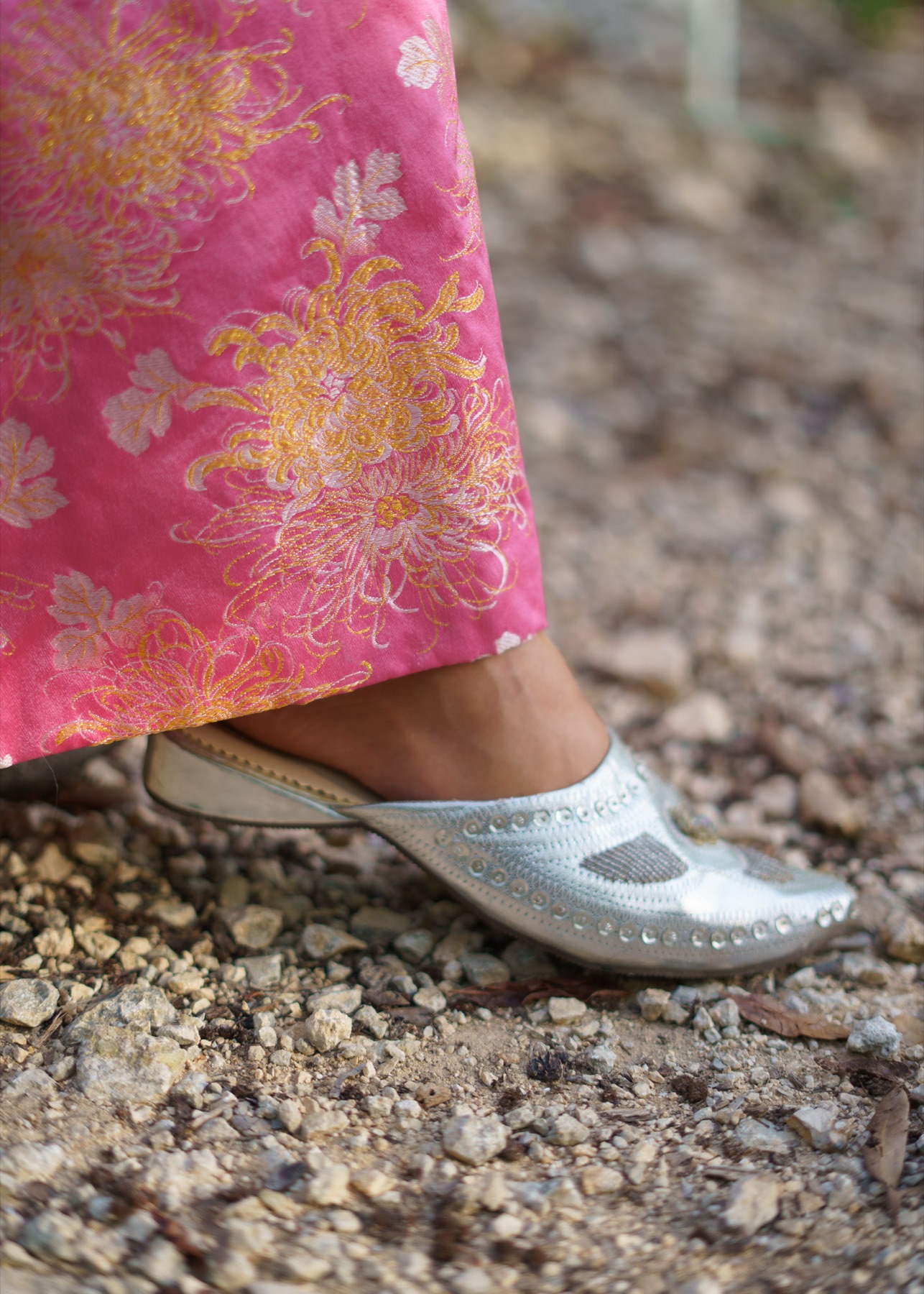 The hem of a pink brocade kimono above a studded silver mule on a gravel path.