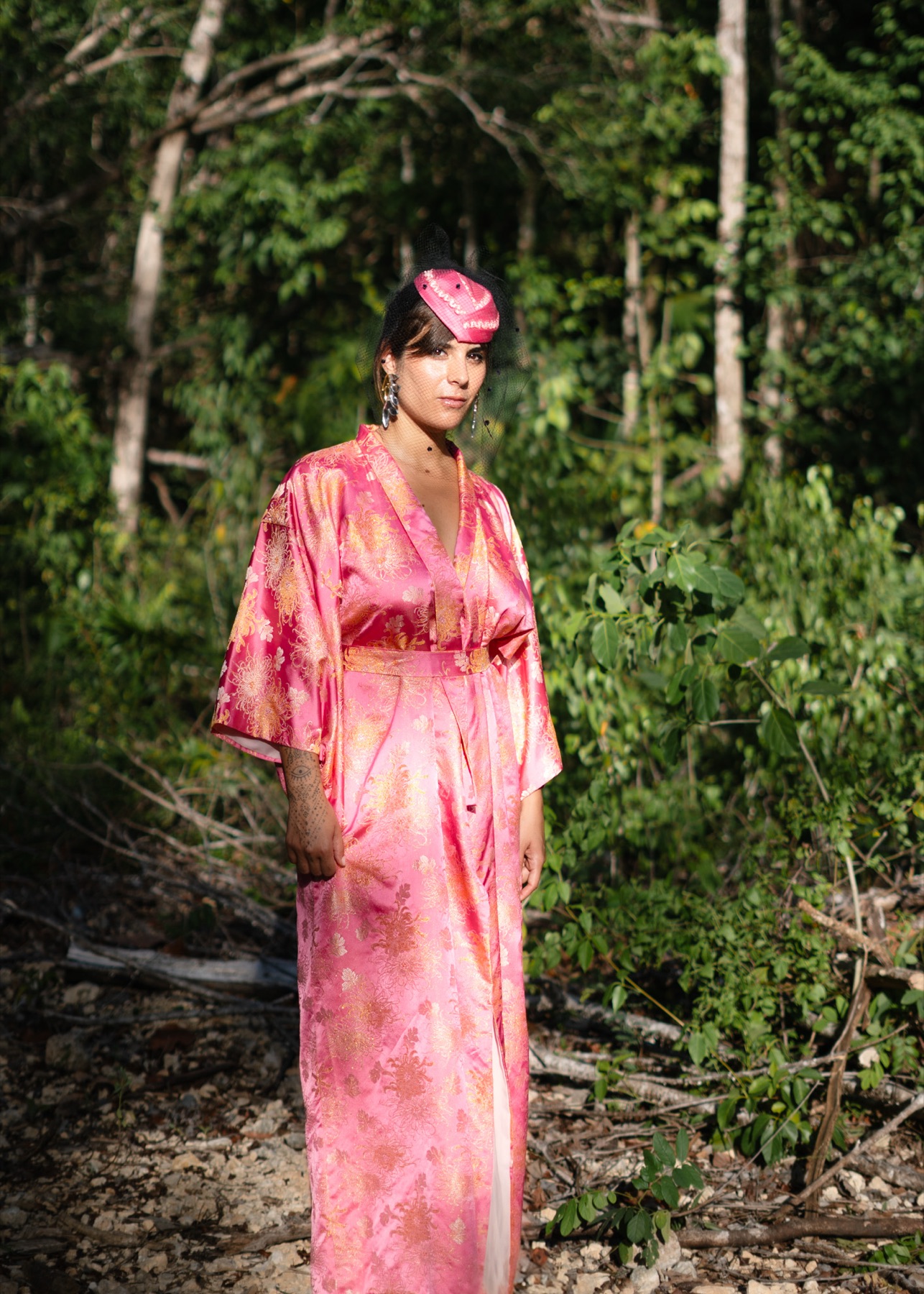 A woman in a pink and gold brocade kimono stands full length in cleared jungle, veiled pillbox cap on her head.