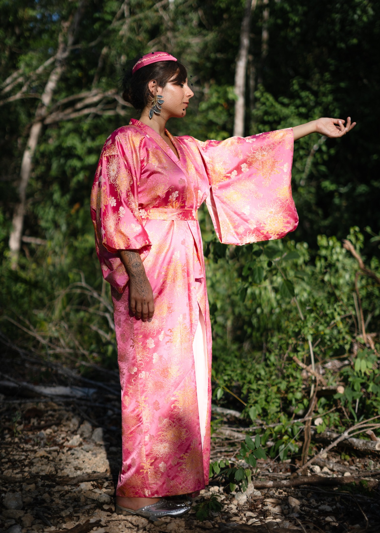 A woman in a pink brocade kimono extends one arm, sleeve falling open, jungle behind her.
