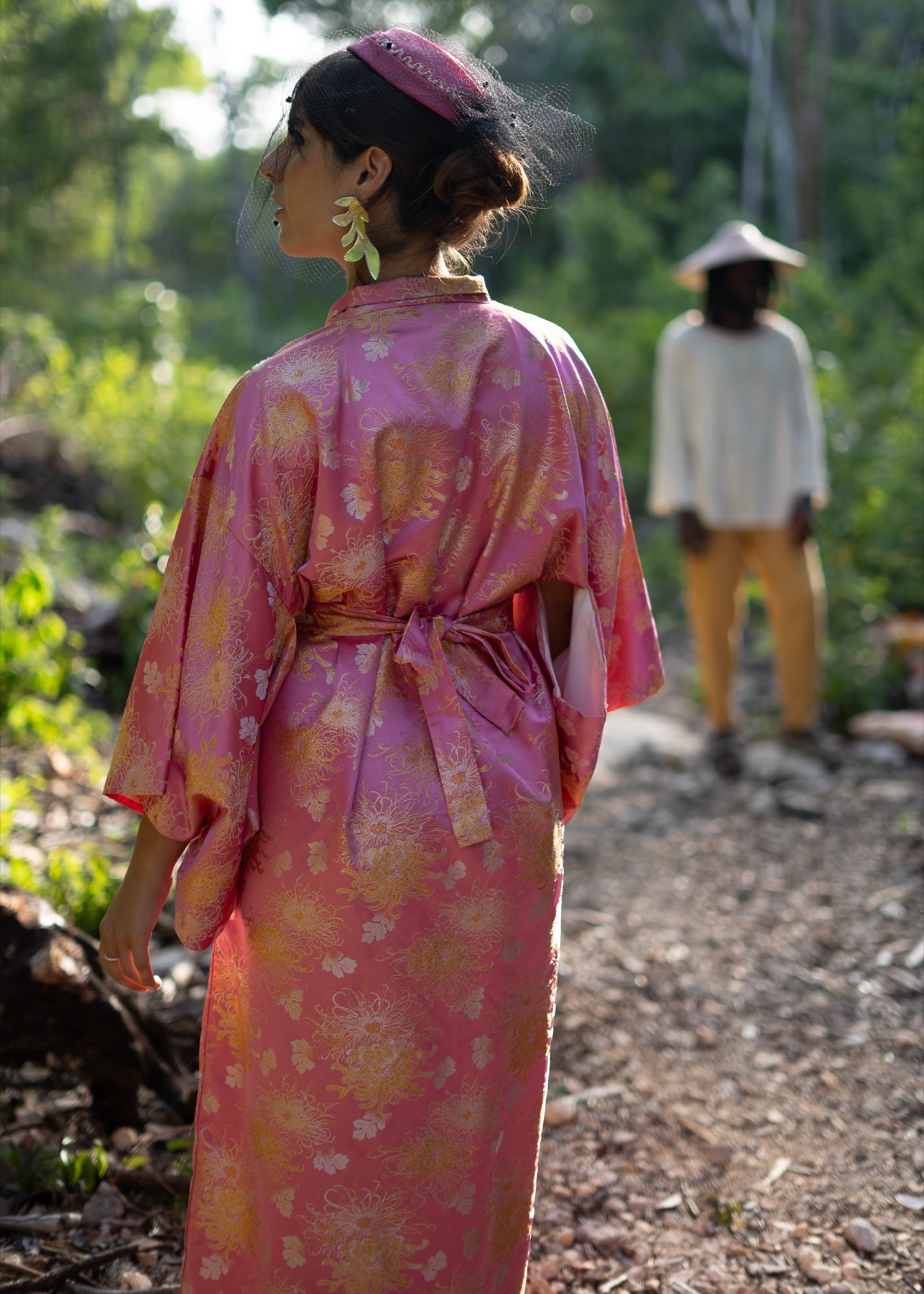A woman in a pink brocade kimono turns away as a figure in a straw hat walks off down the path behind her.