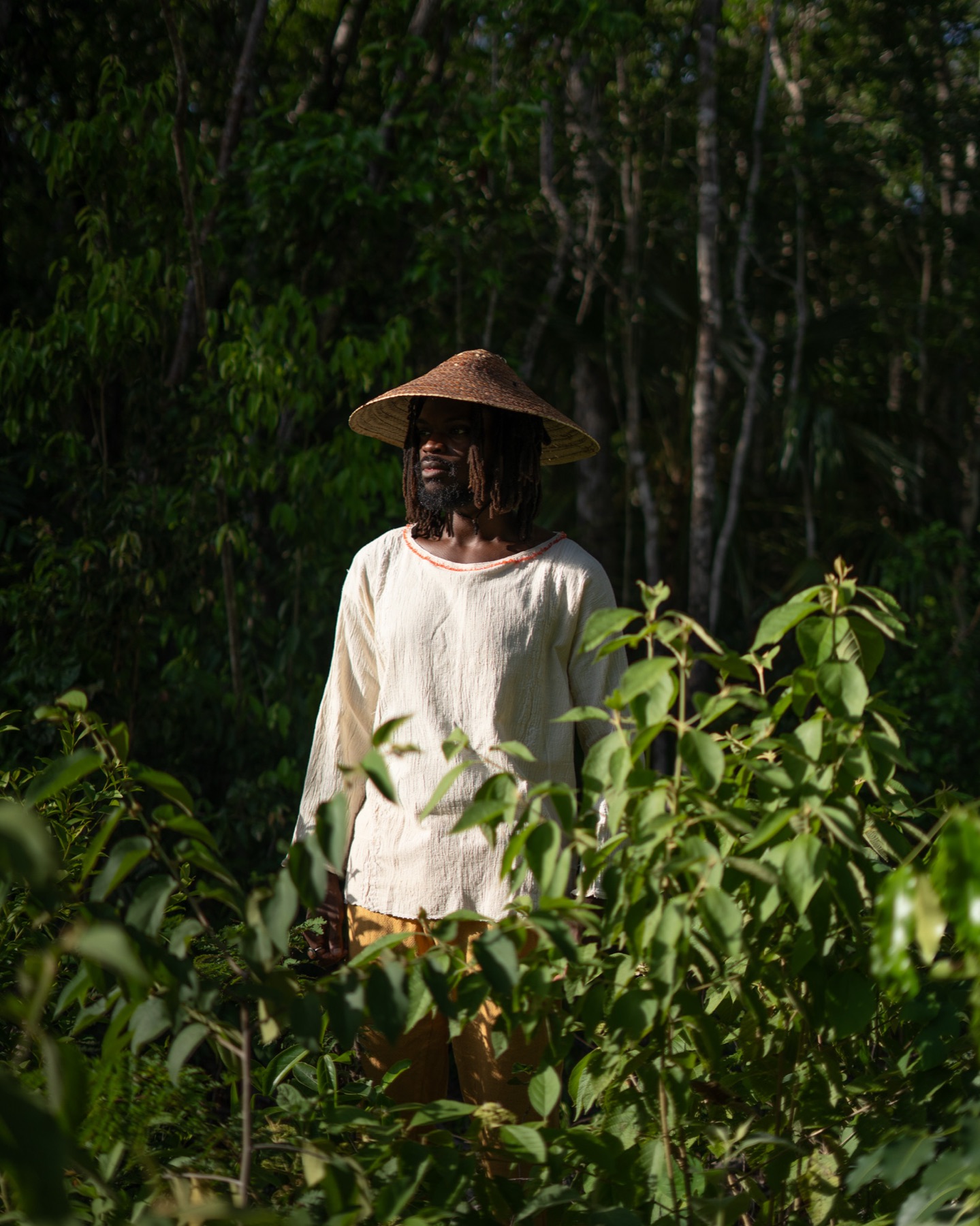 A man in a straw conical hat and cream shirt stands half hidden behind bright jungle foliage.