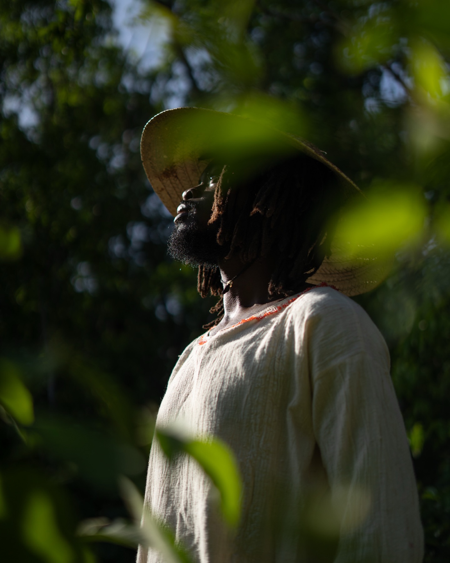 Profile of a man in a straw hat and cream shirt seen through out-of-focus leaves.