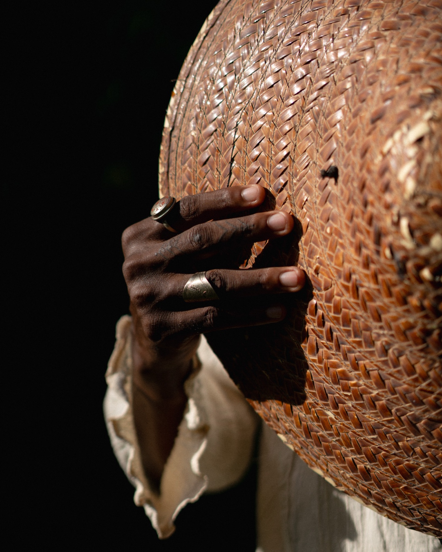 A hand with two silver rings grips the woven brim of a straw hat against a black ground.