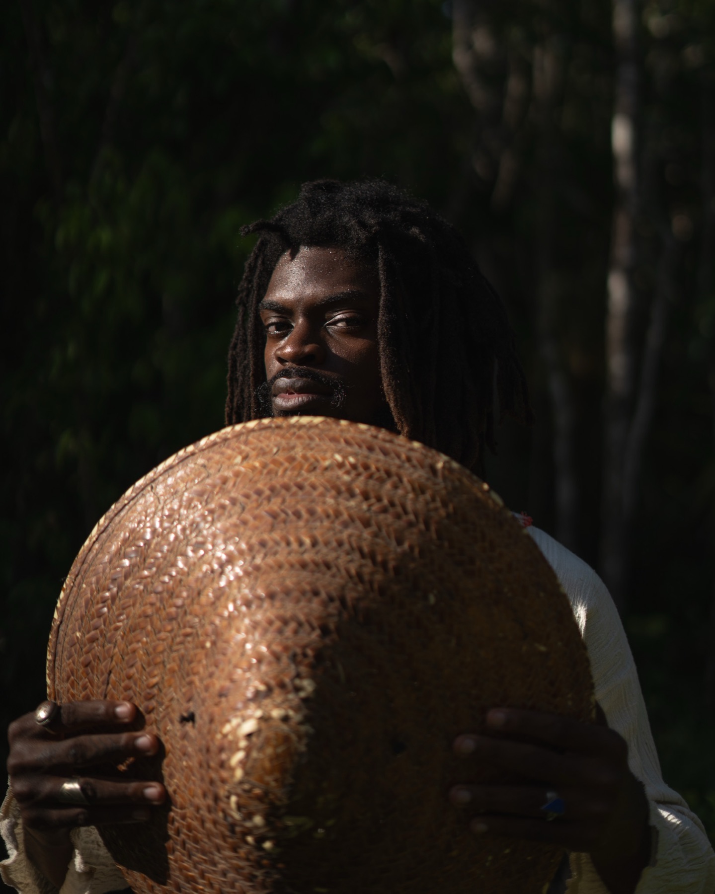 A man holds a woven straw hat against his chest with both hands, rings on his fingers, dark greenery behind.