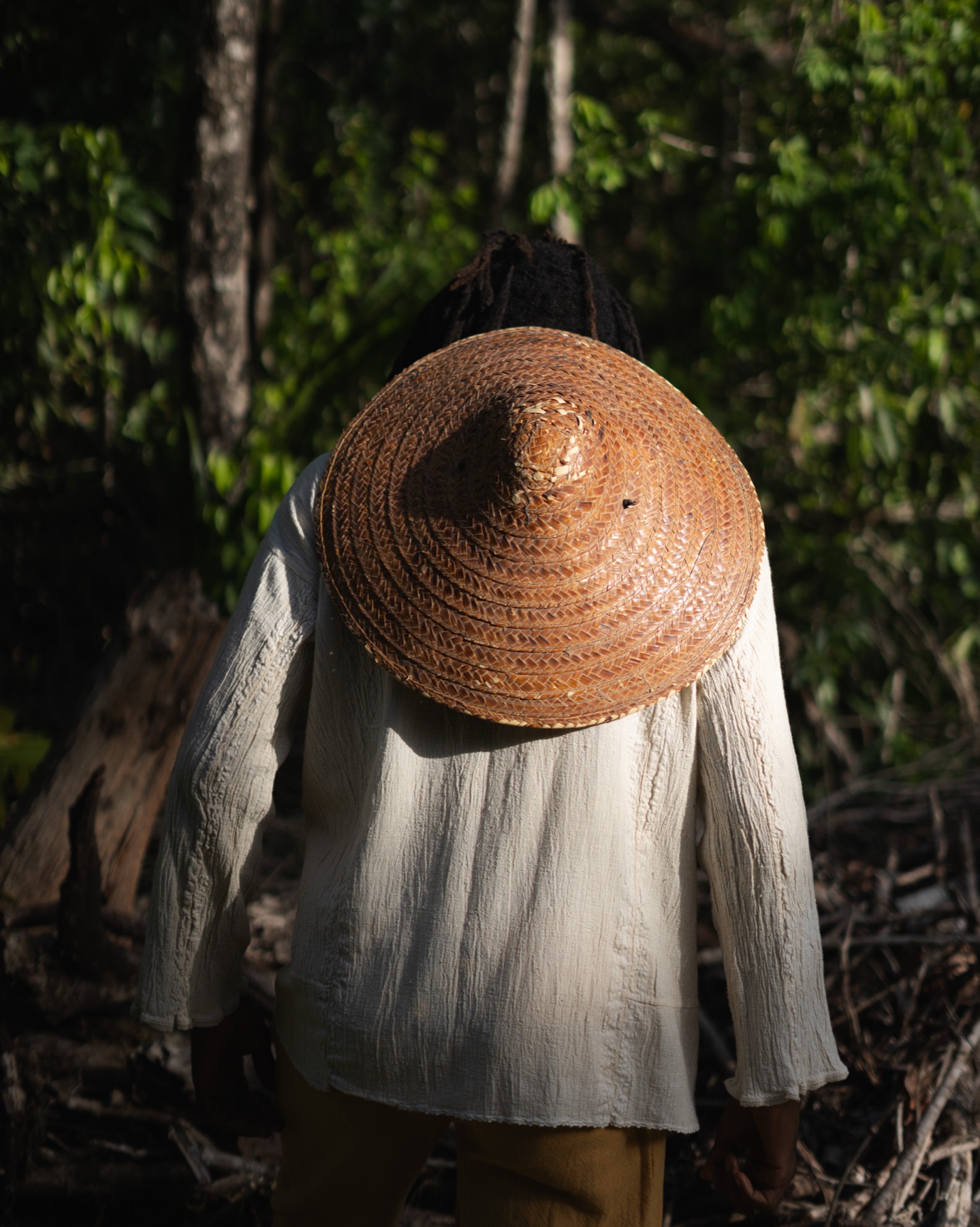 A man seen from behind, straw hat slung on his back over a loose cream shirt, facing into the jungle.