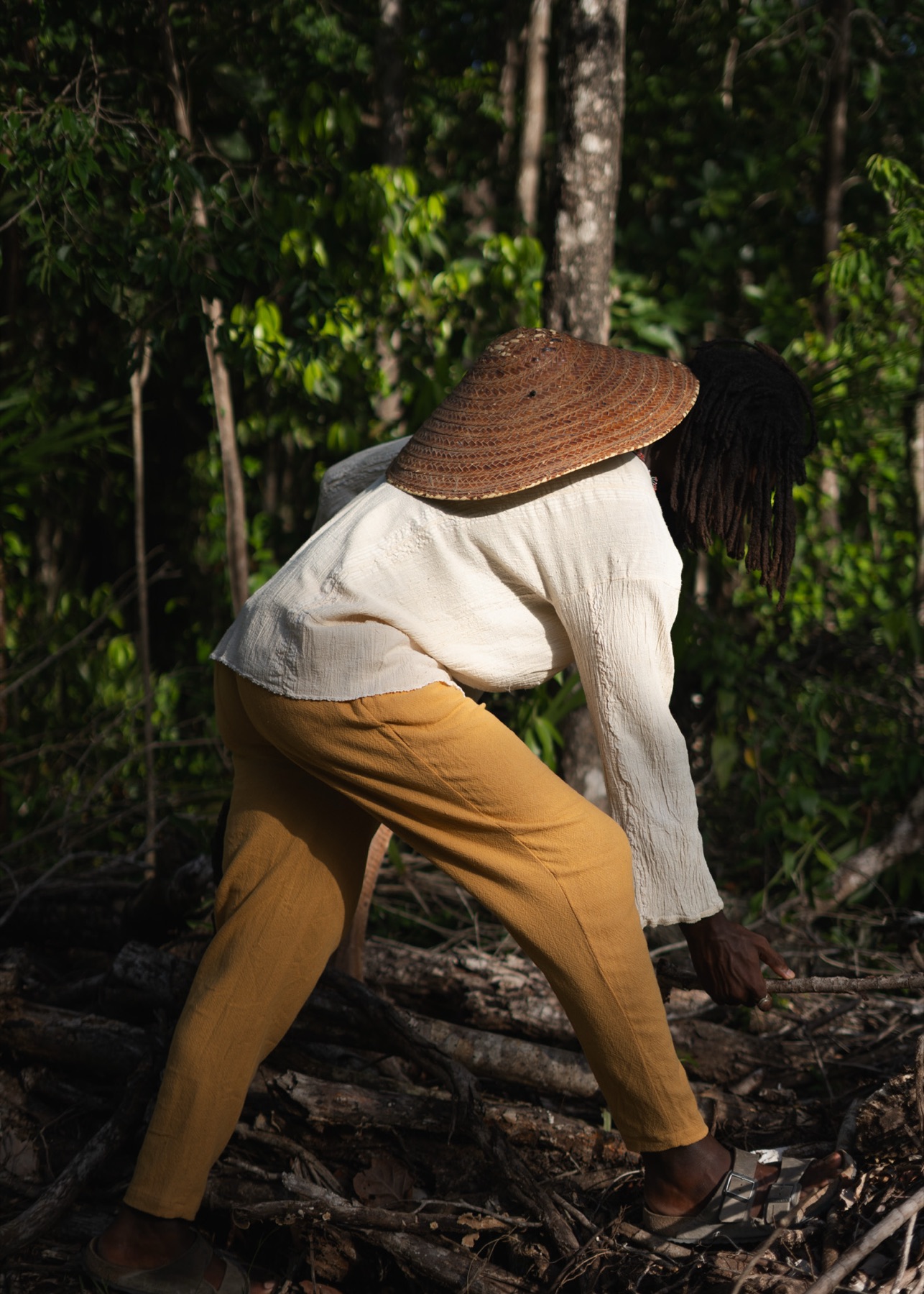 A man in a straw hat bends forward across fallen branches, cream shirt and ochre trousers in motion.