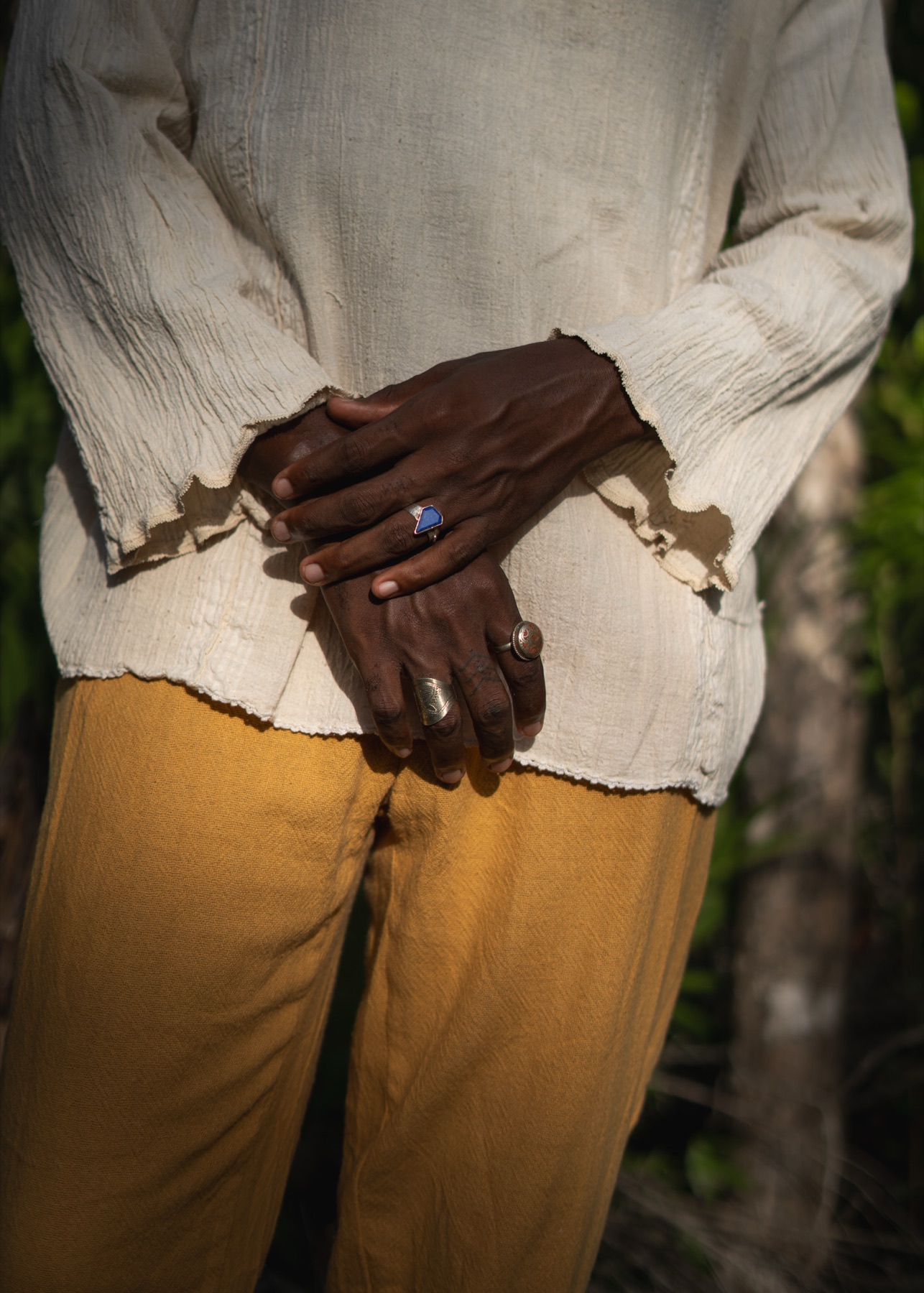 Two hands clasped at the waist, blue stone and silver rings, between a cream shirt and ochre trousers.
