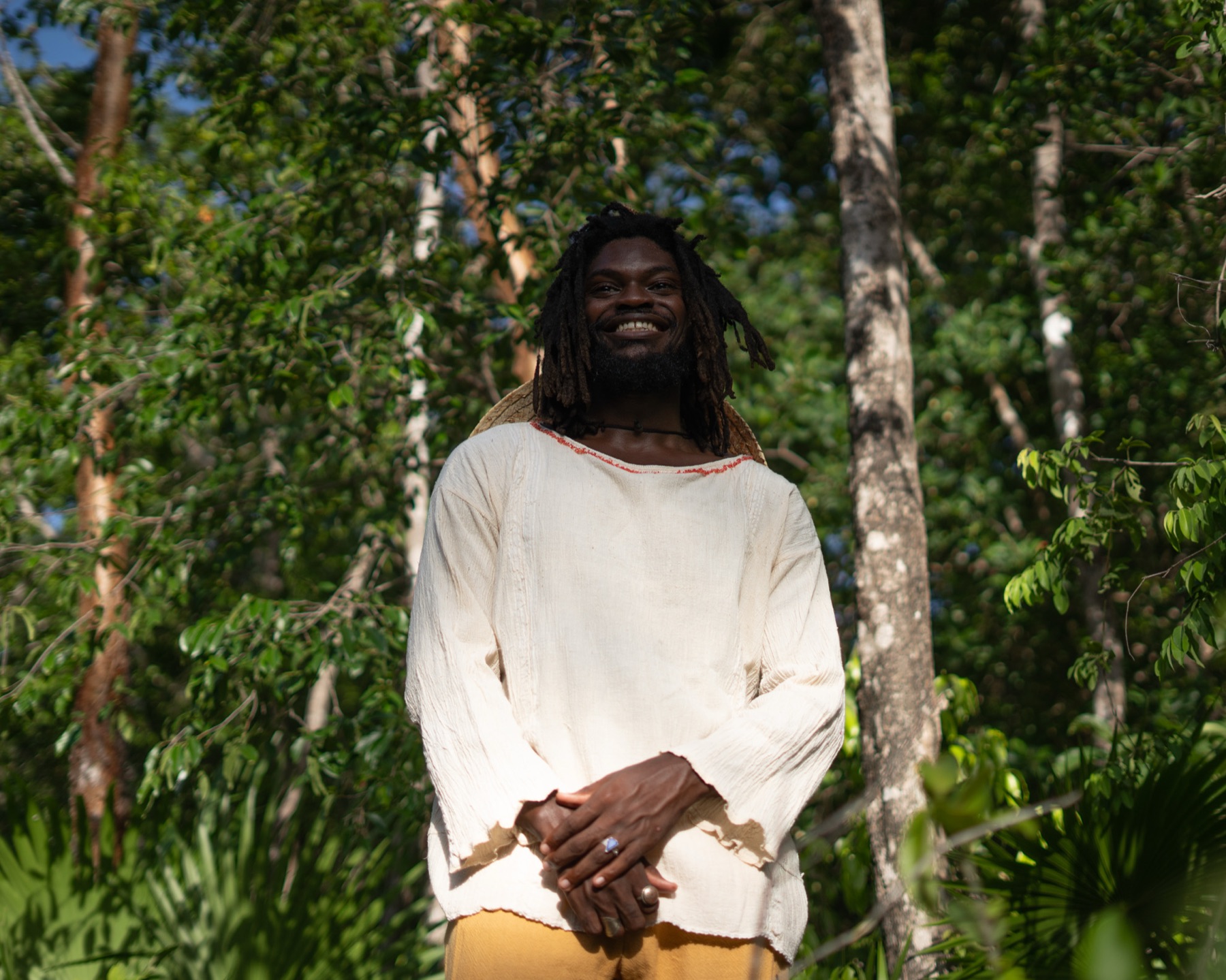 A man smiles broadly with hands clasped, cream shirt and straw hat, sunlit jungle behind him.