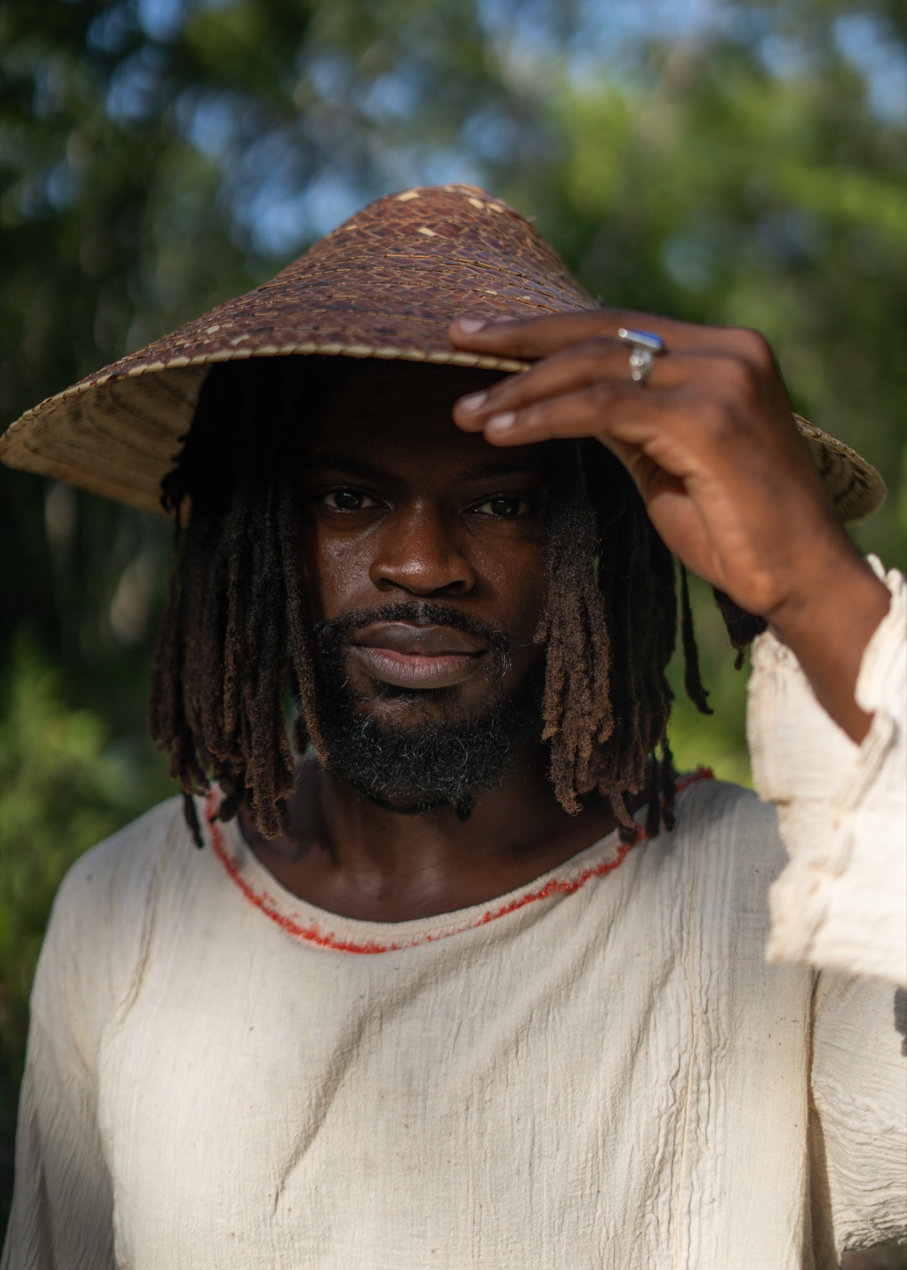 A man tilts the brim of his straw hat, cream shirt with coral trim at the neck, jungle light behind.
