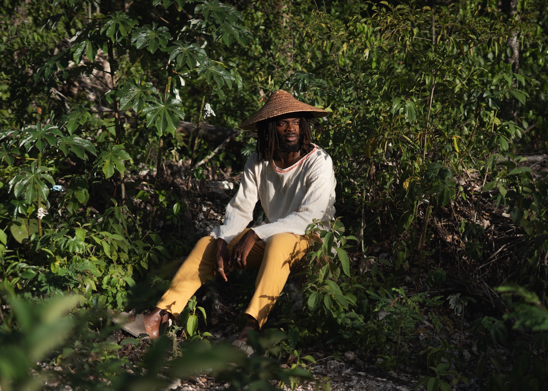 A man sits on limestone rock in a straw hat, cream shirt and ochre trousers, surrounded by low jungle growth.