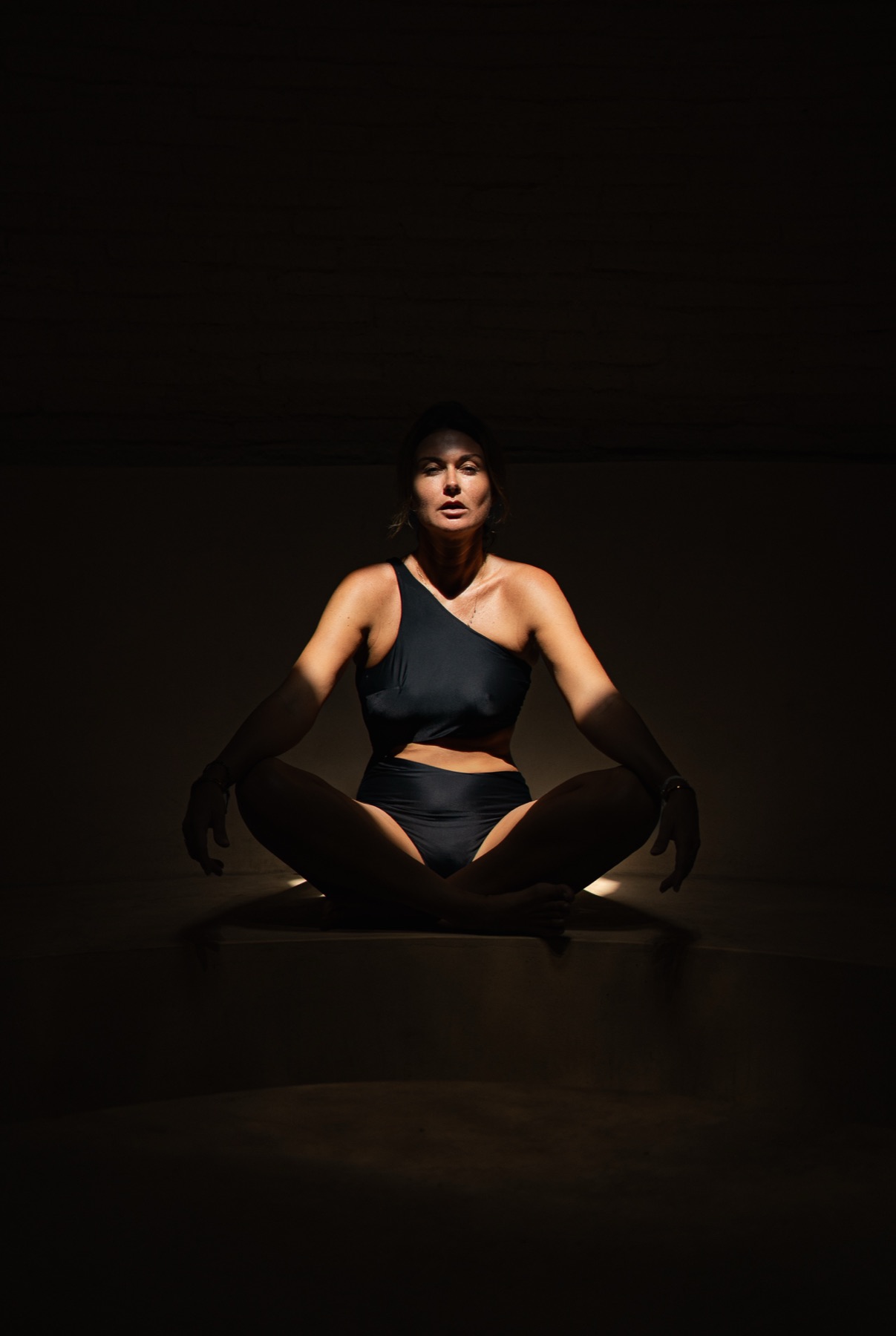 A woman in a one-shoulder black bodysuit and sheer gloves sits cross-legged in a dark studio, lit from below.