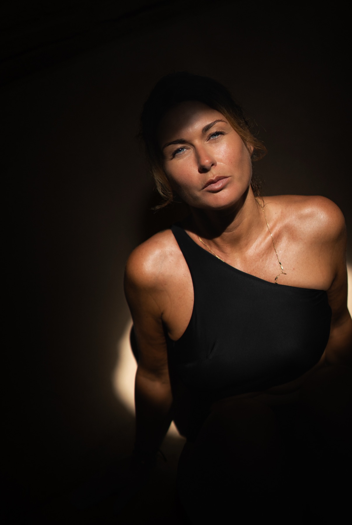 Low-key portrait of a woman in a one-shoulder black top leaning into a single shaft of light.