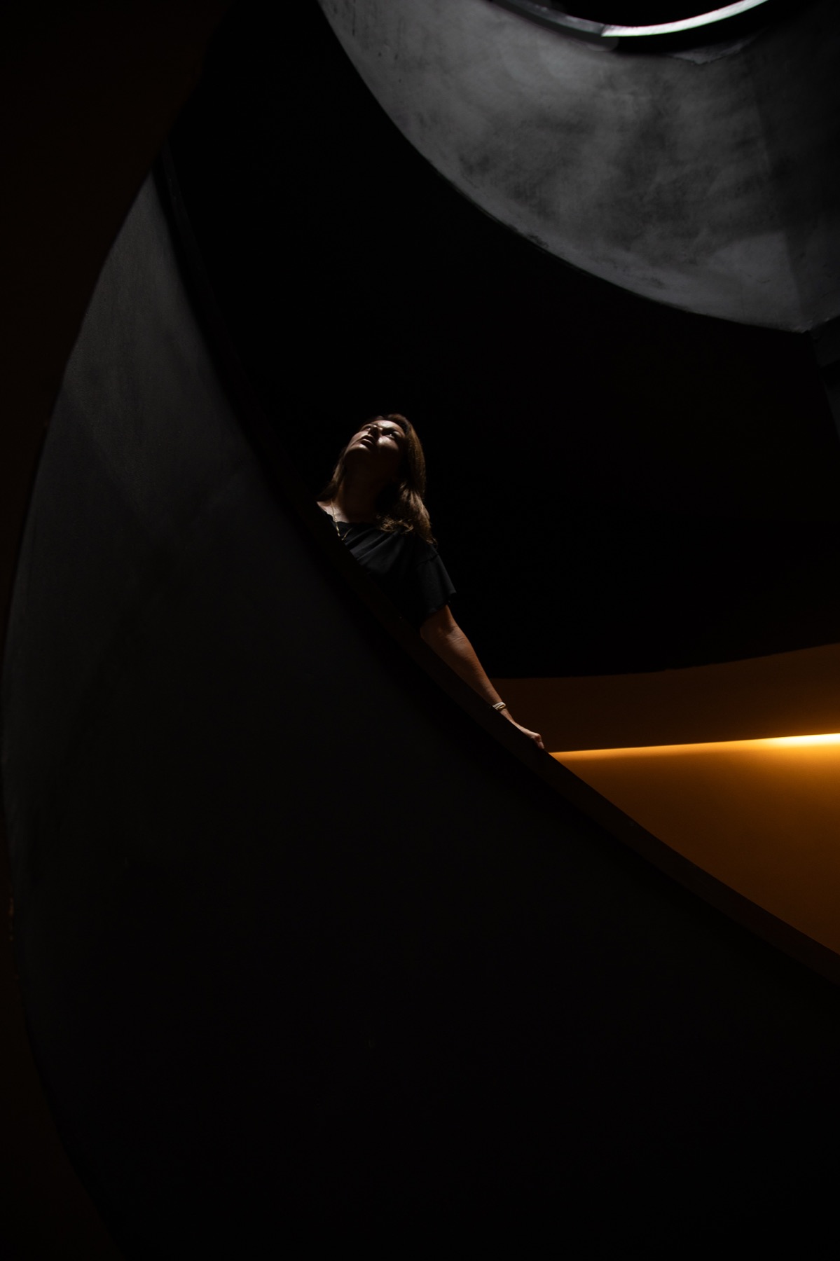 A woman looks up from inside a dark curved concrete stairwell, a band of amber light behind her.