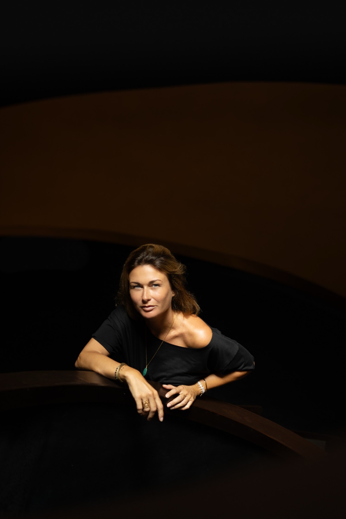 A woman in a black off-shoulder top leans on a curved dark wall wearing a turquoise pendant.