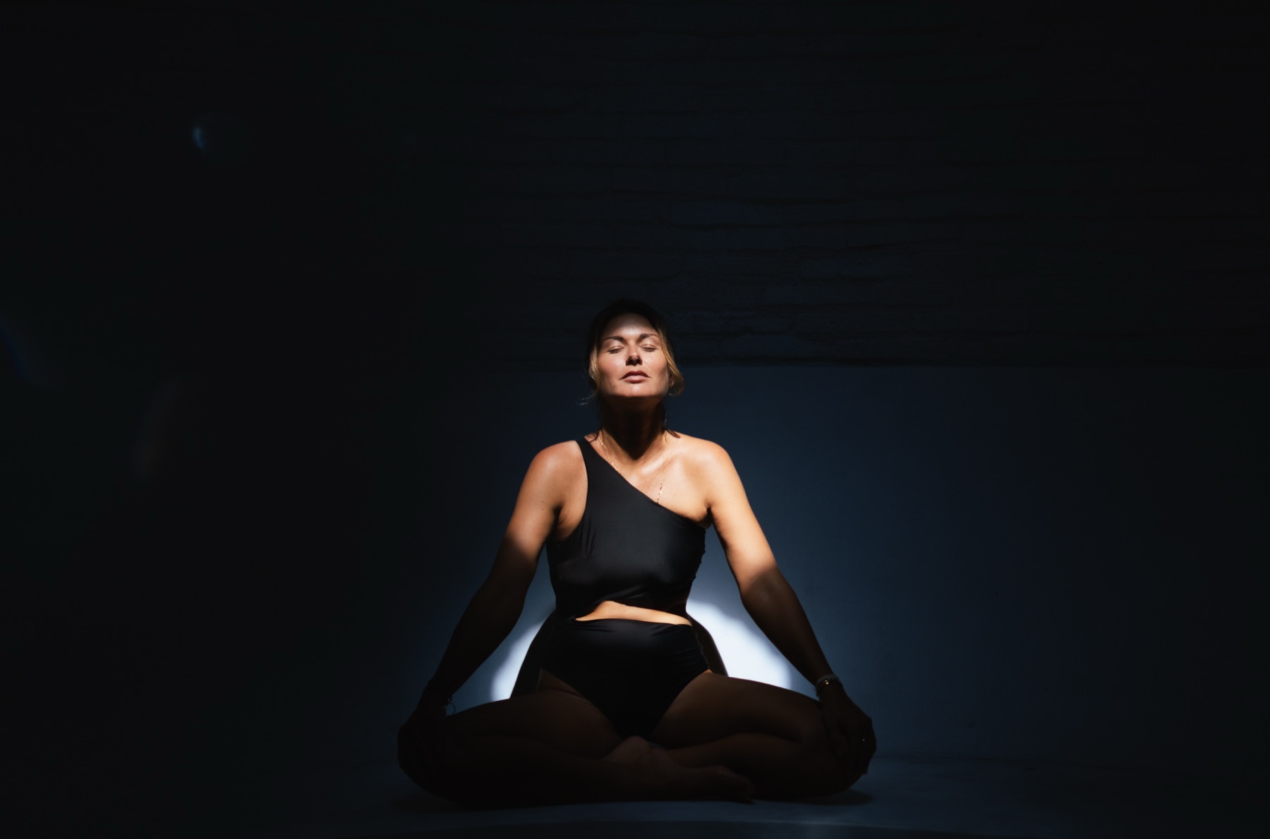 A woman sits cross-legged in a black bodysuit against a dark wall, eyes closed, light pooling behind her.