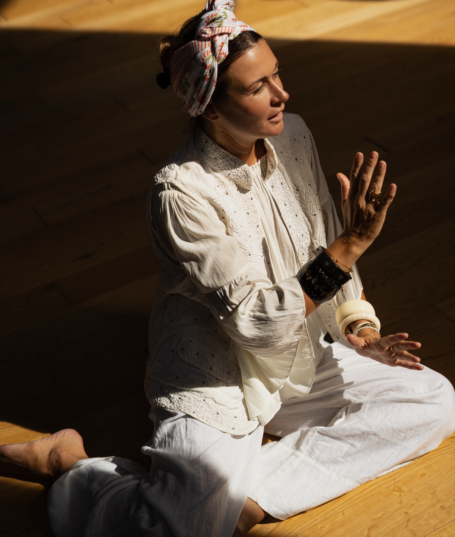 A woman in a white eyelet blouse and floral headscarf sits on a wooden floor, hands raised as she speaks.