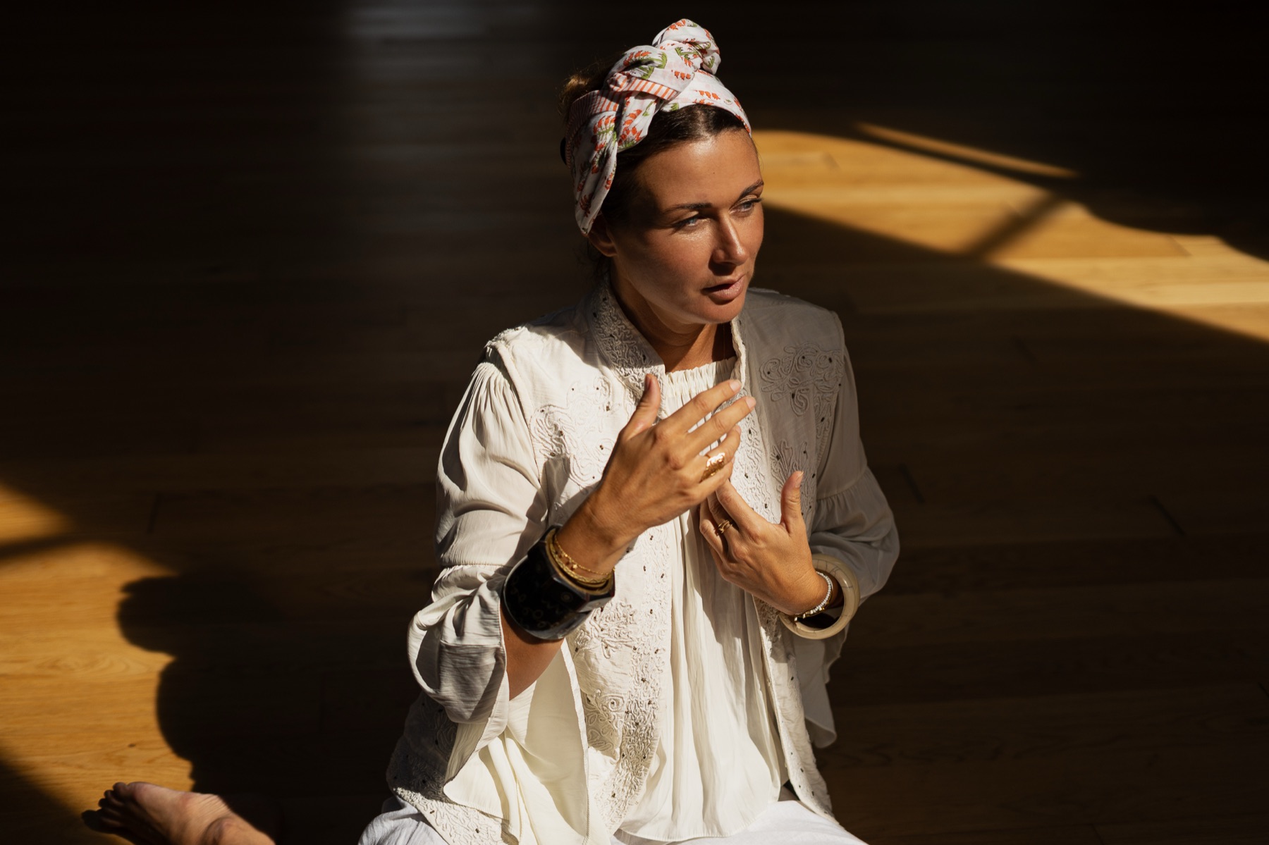 A woman in white linen and a floral headscarf presses a hand to her chest in hard window light.