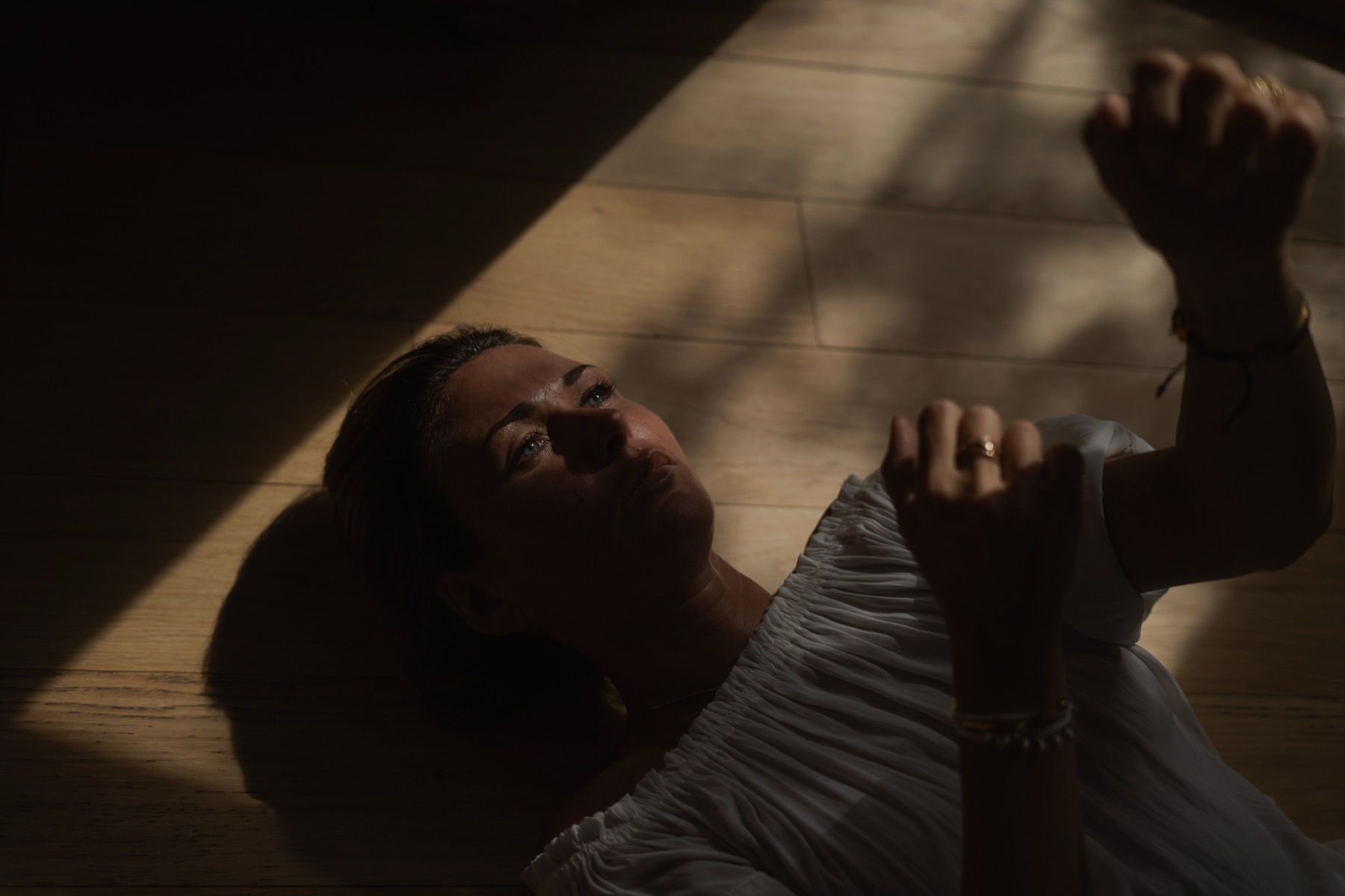 A woman lies on a wooden floor in white cotton, hands raised into a shaft of afternoon light.