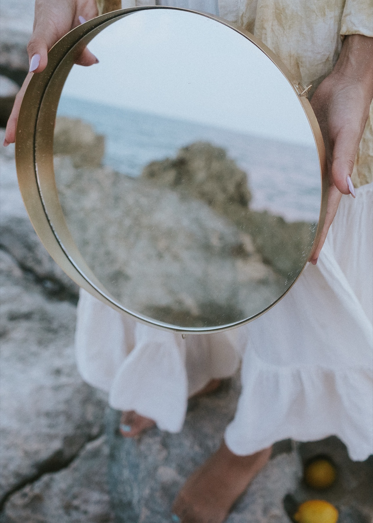 Two hands hold a round brass mirror reflecting the rocks and sea, white linen skirt behind.