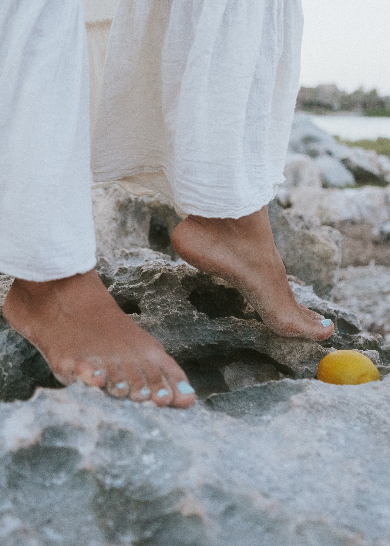 Bare feet with pale blue polish on limestone rock beside a lemon, white linen skirt above.