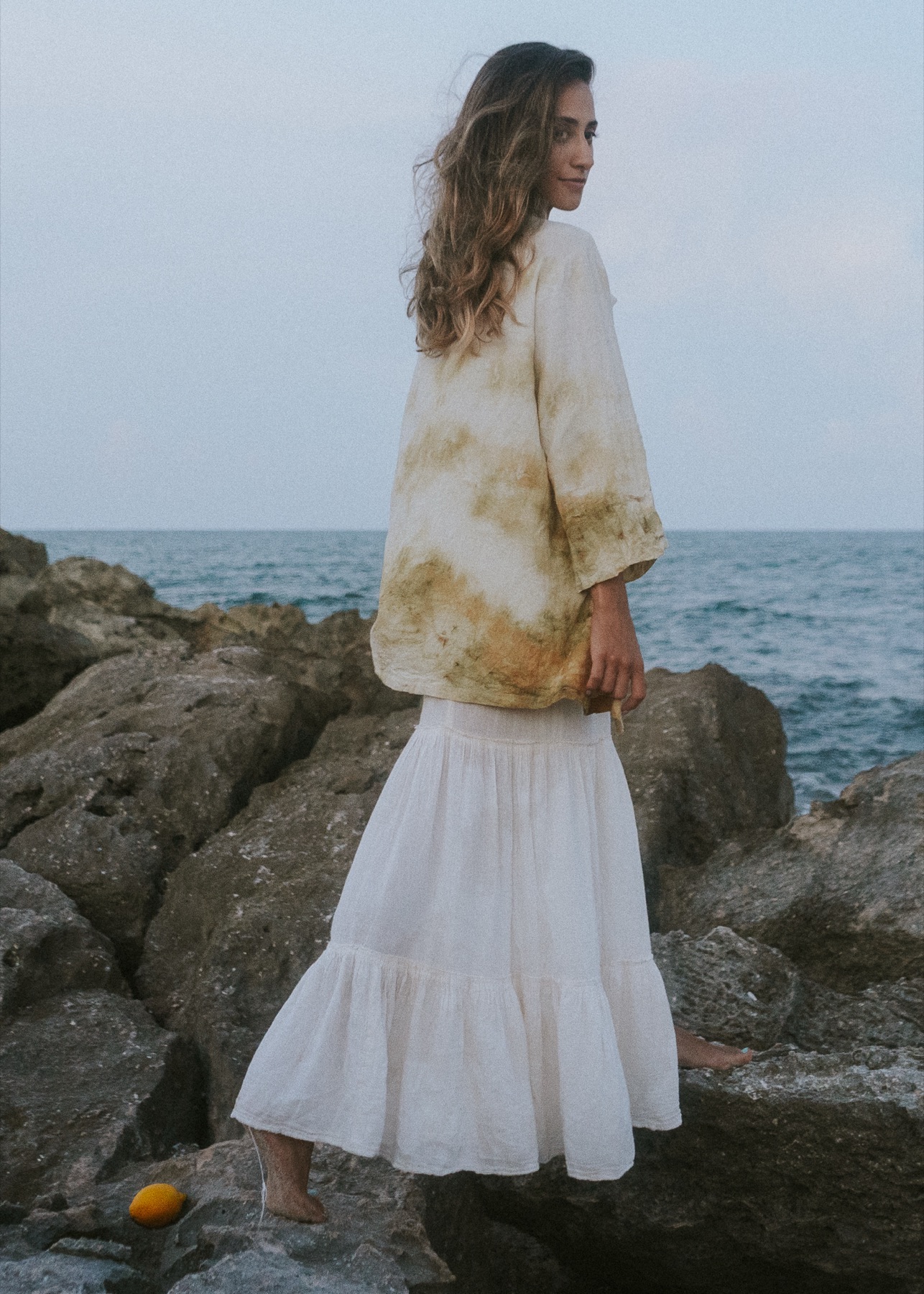 A woman in a botanically dyed jacket and long white skirt looks back over her shoulder on limestone rocks by the sea.