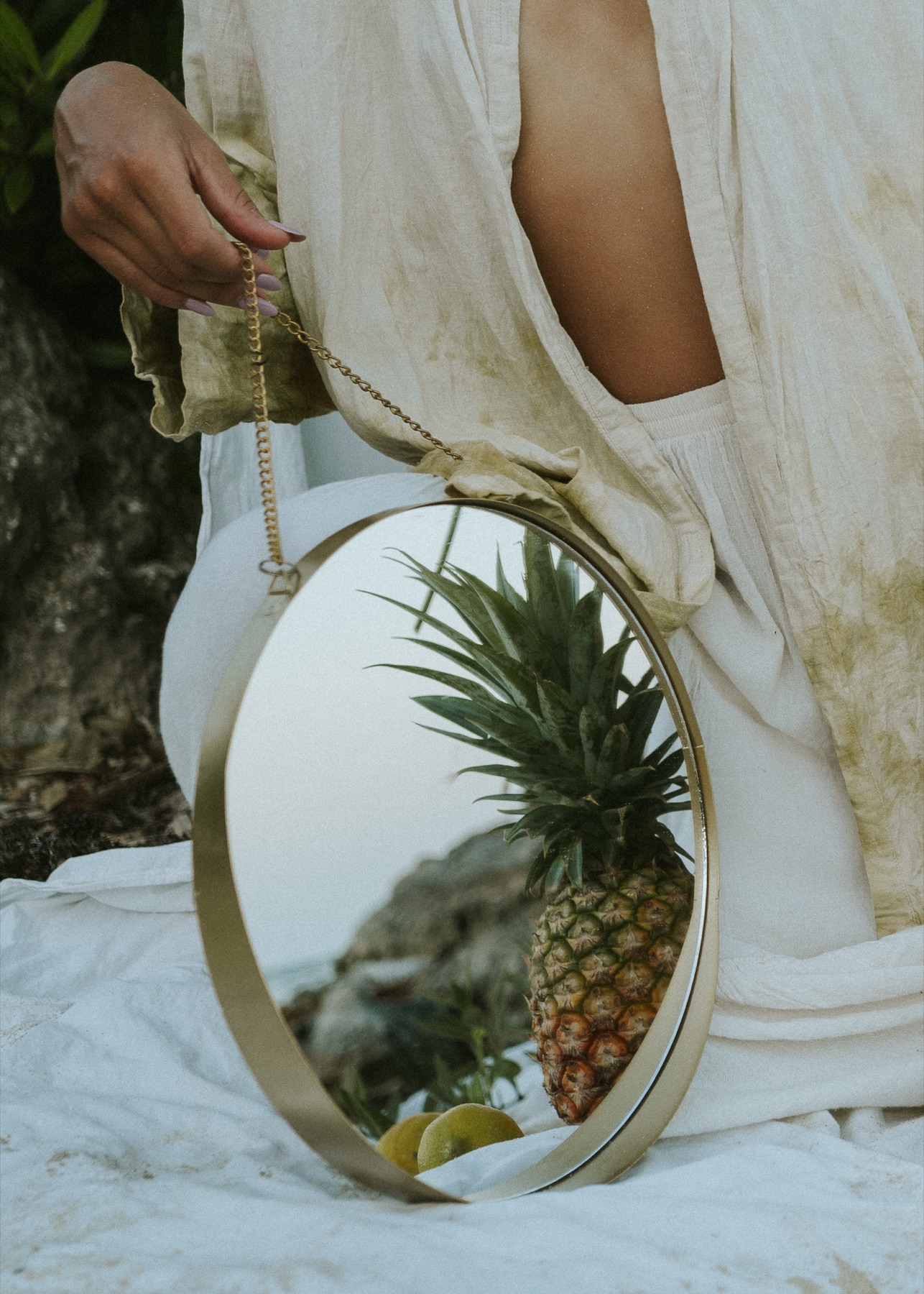 A hand holds a round brass mirror beside a pineapple and lemons on white cloth, reflecting the fruit.